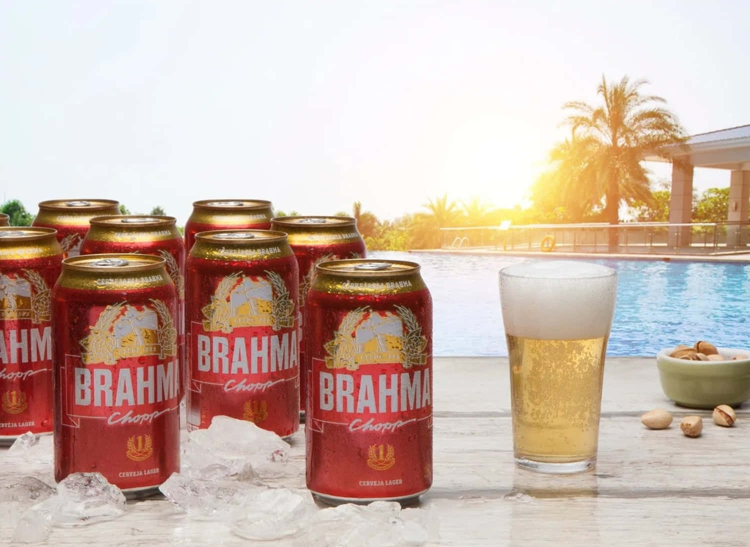 巴西布拉馬Chopp啤酒罐泳池畔 壁紙 — 紅色Brahma啤酒罐和一個有泡的玻璃杯放在泳池邊的冰上，背景是金色日落和棕櫚樹。