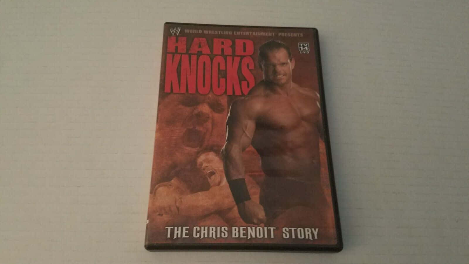Chris Benoit DVD封面 壁紙 — 《Hard Knocks: The Chris Benoit Story》的DVD封面，展示一位肌肉發達的摔角手站在倒下的對手之上，背景是火紅色的。