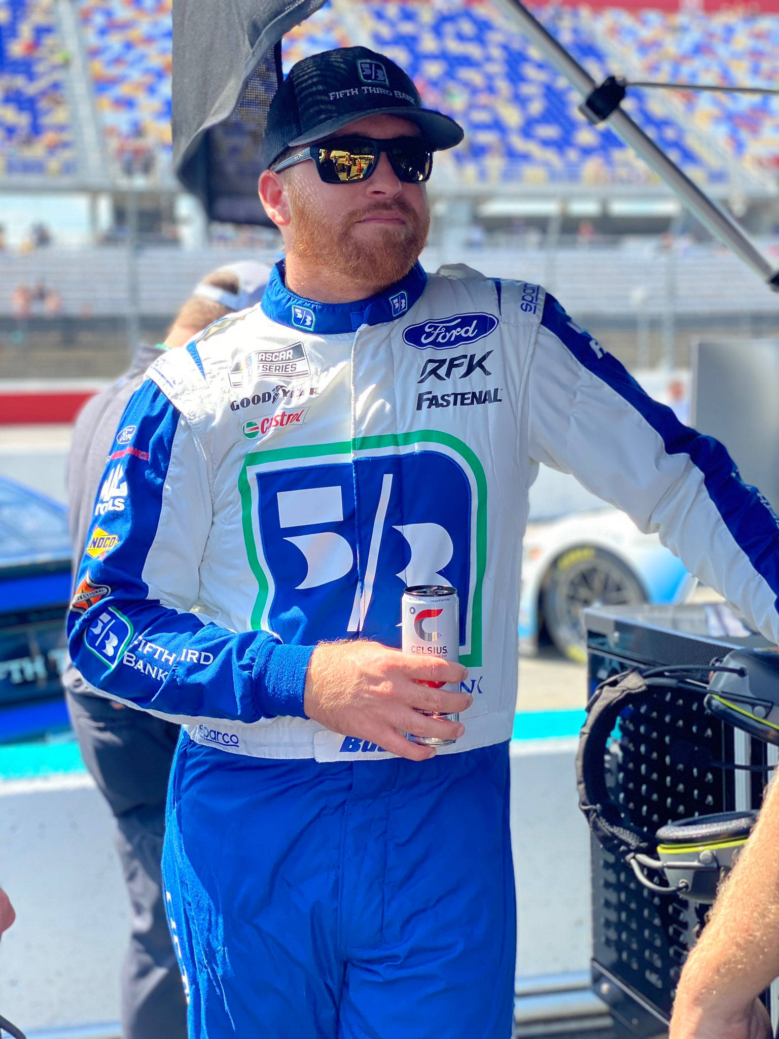 Chris Buescher 放鬆時刻 壁紙 — 一位身穿藍白賽車服的NASCAR車手，在賽道上的遮陽篷下拿著一個罐子。