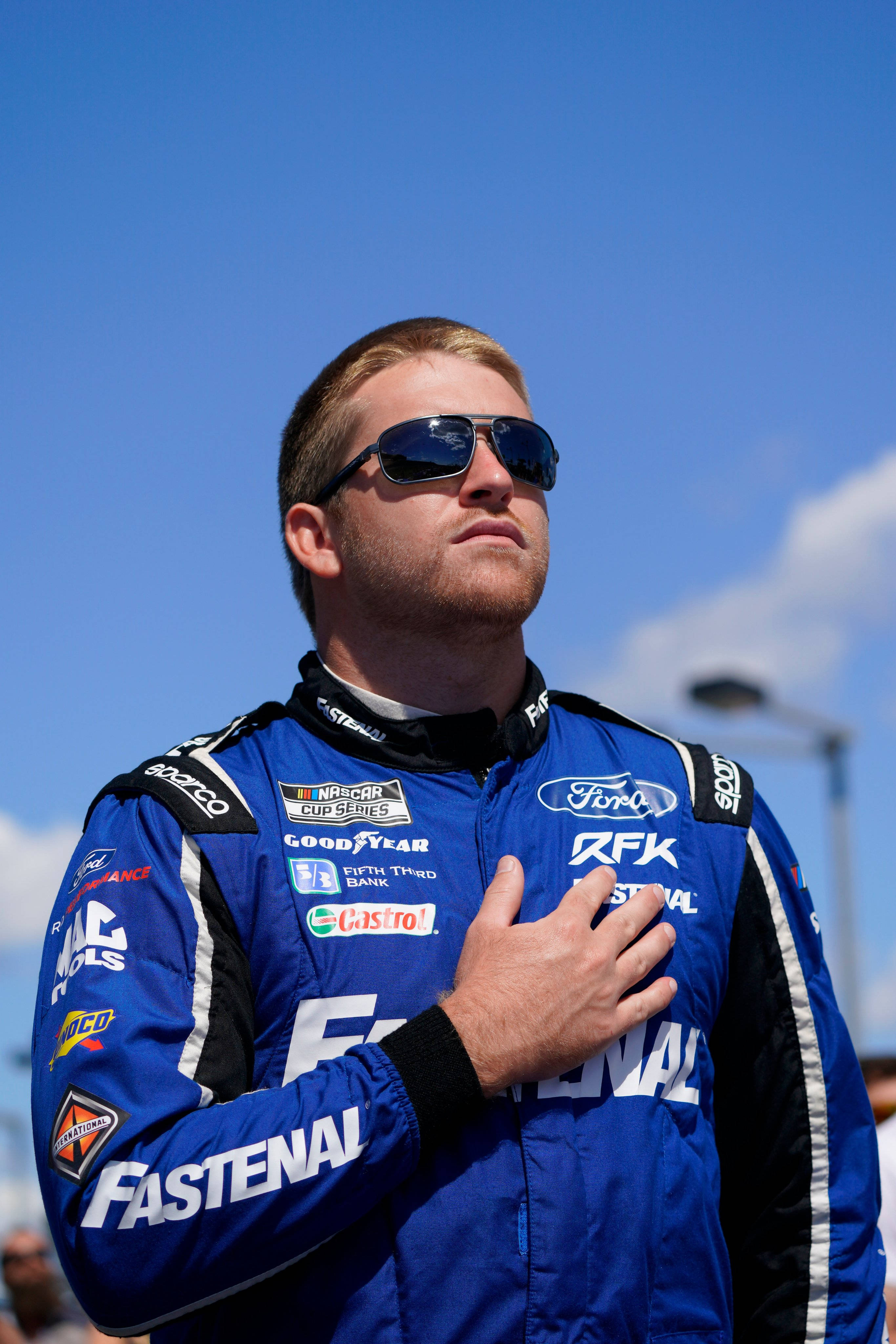 Chris Buescher 手放胸口 壁紙 — 一位身穿藍色賽車服的NASCAR車手在晴朗的藍天下莊嚴地將手放在心口上。