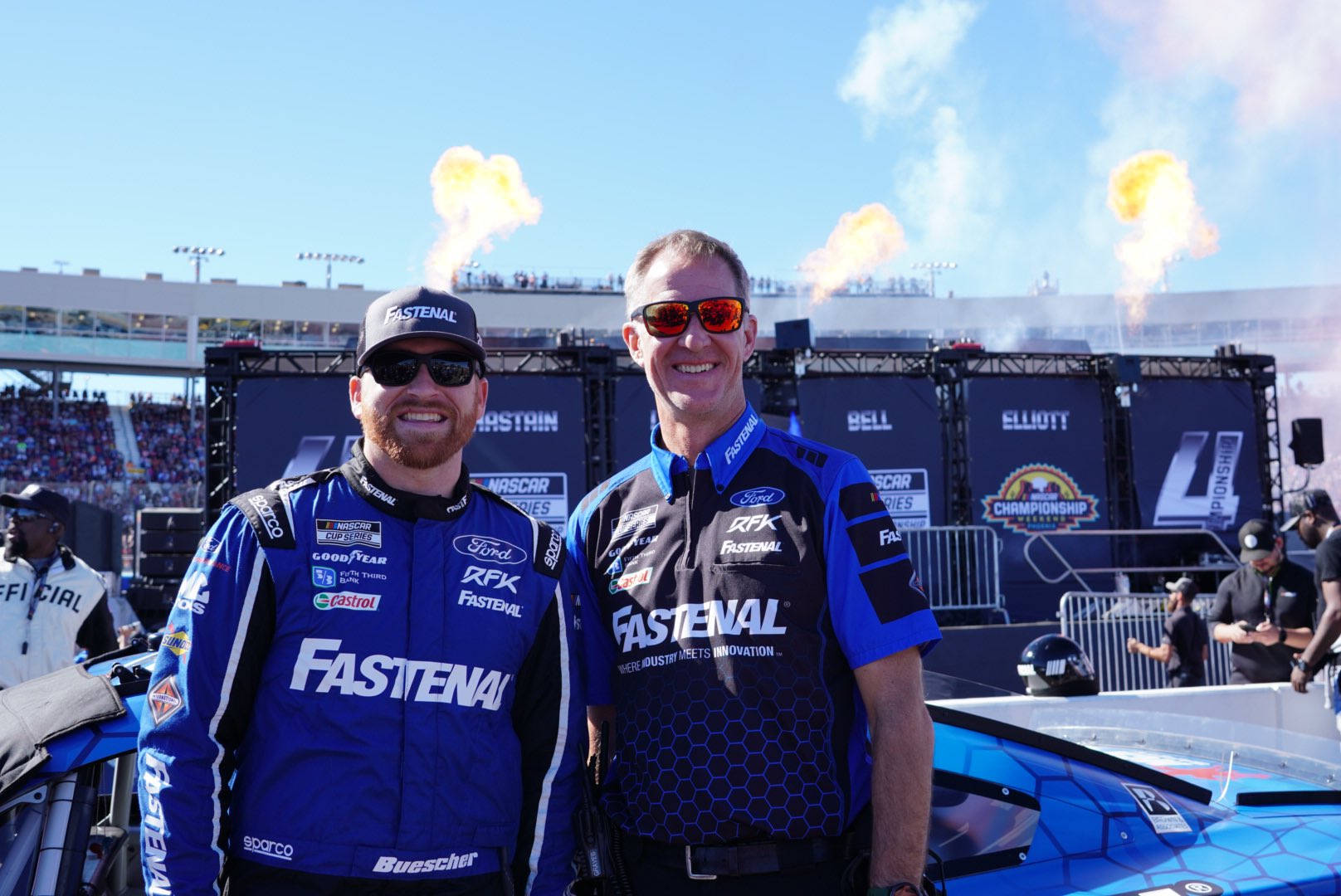 兩名身穿藍色賽車服的NASCAR車手在賽車旁微笑，身後有煙火。