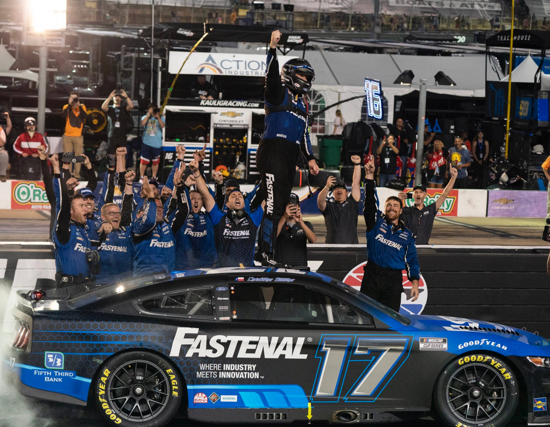 Chris Buescher 賽後歡呼 壁紙 — 一名身穿藍色裝備的NASCAR車手在夜間被他的團隊抬到他的藍黑#17 Fastenal賽車上，舉起拳頭慶祝勝利，背景是歡呼的粉絲。