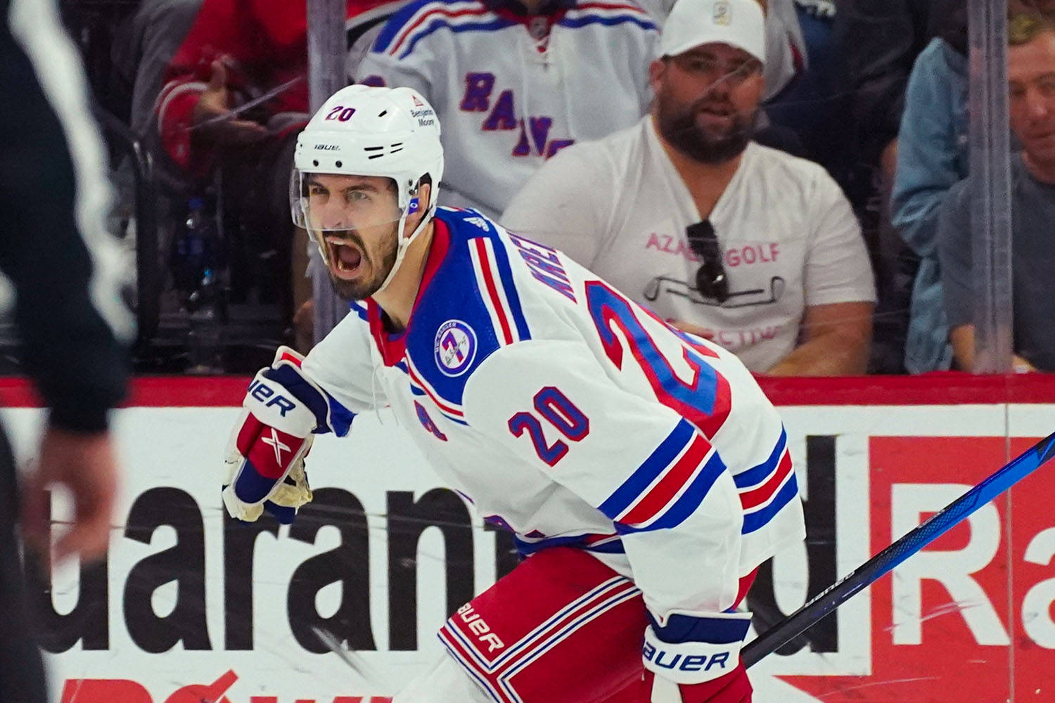 Chris Kreider 冰球鋒線球員 壁紙 — 一名身穿白色、藍色和紅色球衣的冰球運動員在冰上比賽中，背景有球迷，激烈地咆哮。