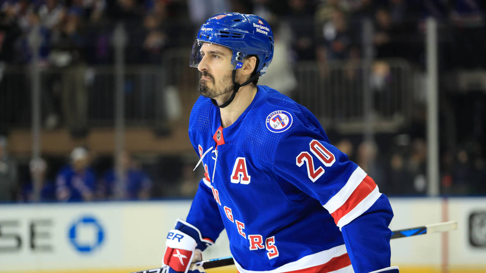 Chris Kreider 美國冰球運動員 壁紙 — 一位專注的曲棍球運動員身穿藍色流浪者隊球衣，號碼20，在明亮的場館燈光下於比賽中在冰上滑行。