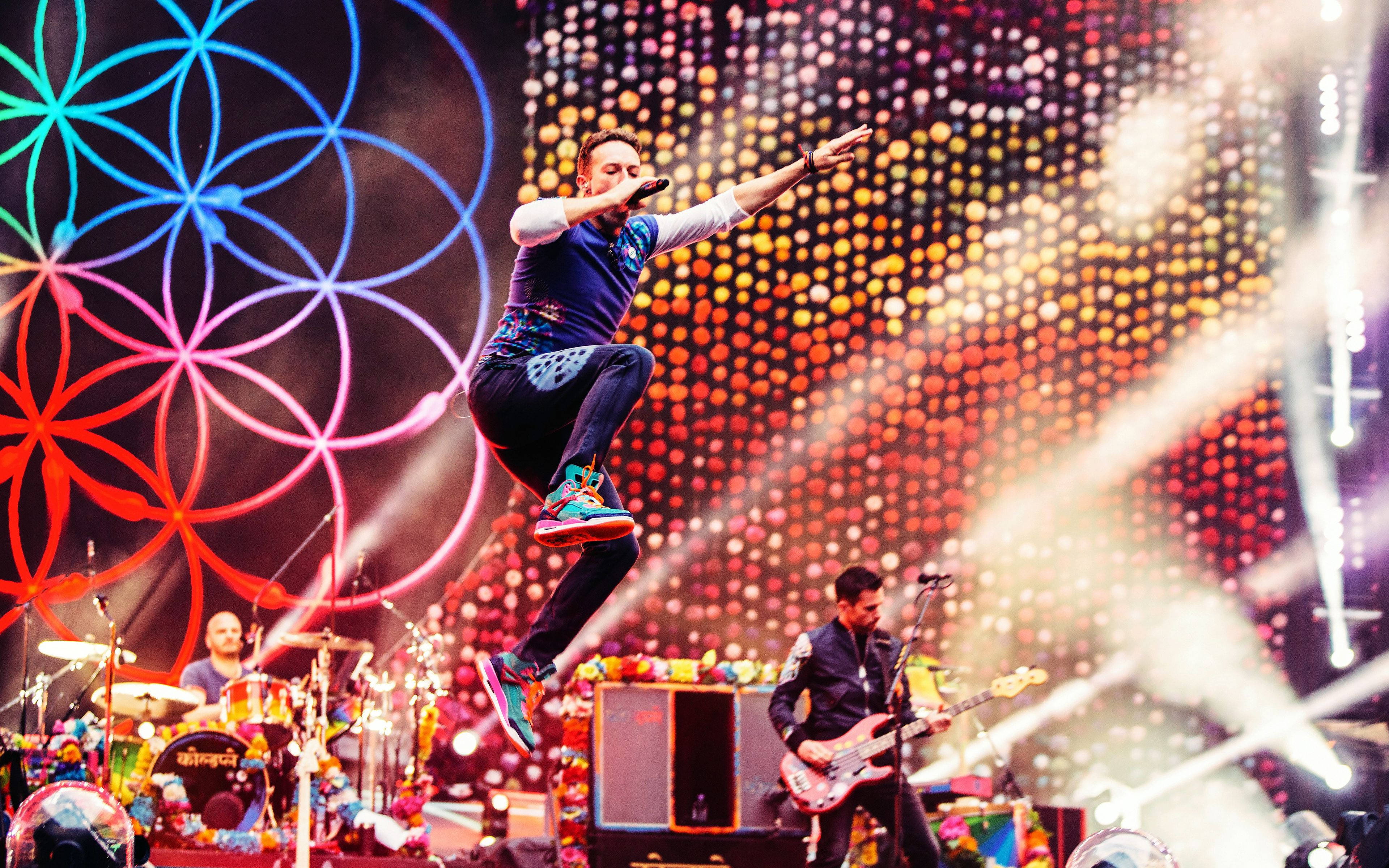Chris Martin A Head Full of Dreams 巡迴演出 壁紙 — 一位歌手在充滿活力的舞台上躍起，舞台上有色彩繽紛的幾何燈光和閃爍的背景。