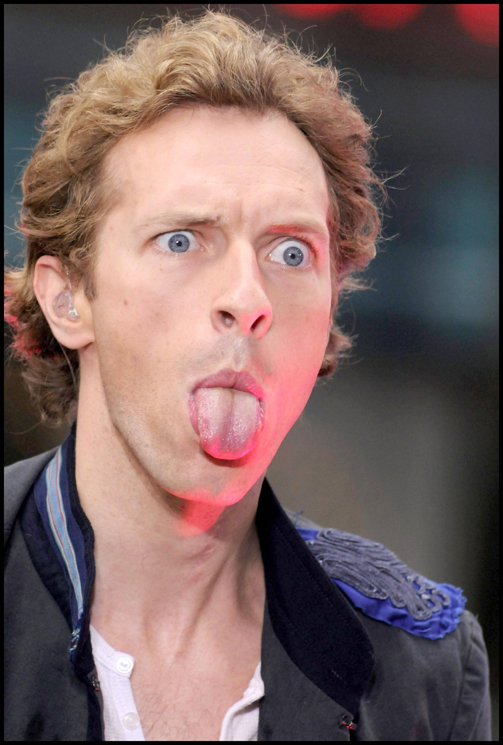 Chris Martin 搞笑表情 壁紙 — 一個捲髮藍眼的男人在舞台燈光下做出頑皮的表情伸出舌頭。