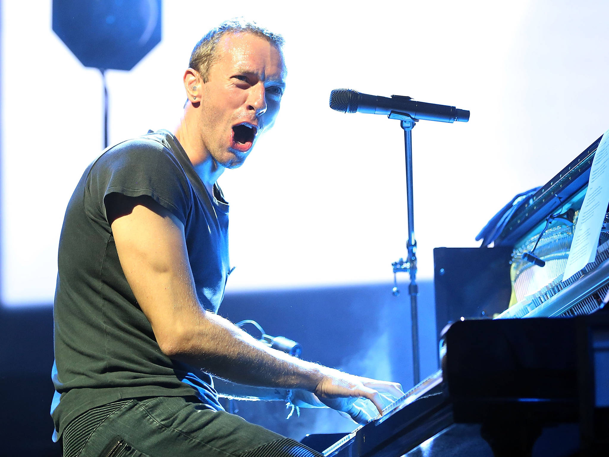 Chris Martin 格拉斯頓伯里音樂節 壁紙 — 一位男歌手在明亮的藍色舞台燈光下，熱情地演奏三角鋼琴並表演。