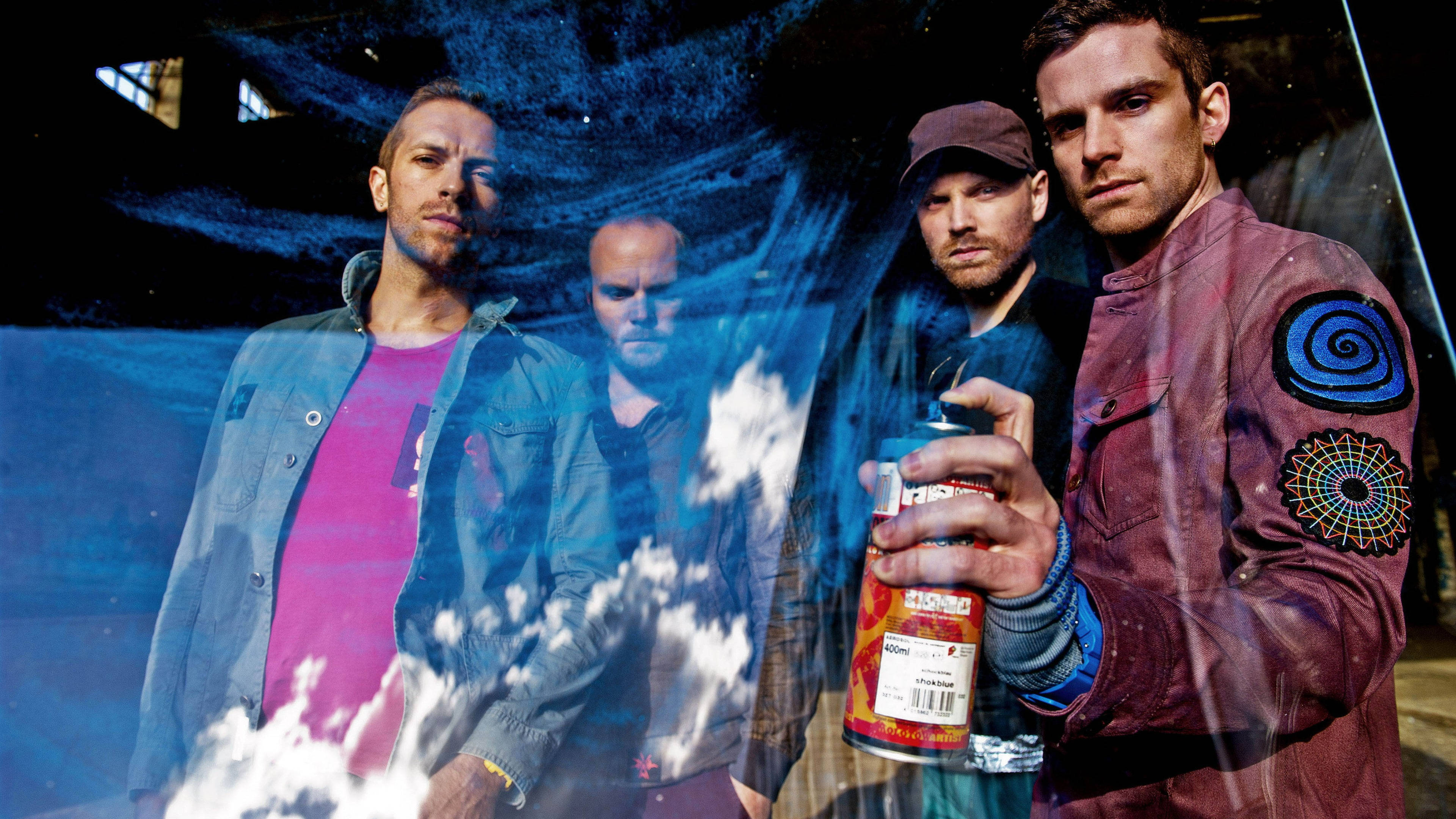Chris Martin 與 Coldplay 乐队成员 壁紙 — 四個穿著都市服裝的男人站在黑暗背景前，帶有藍色噴漆效果，其中一人手持噴漆罐。