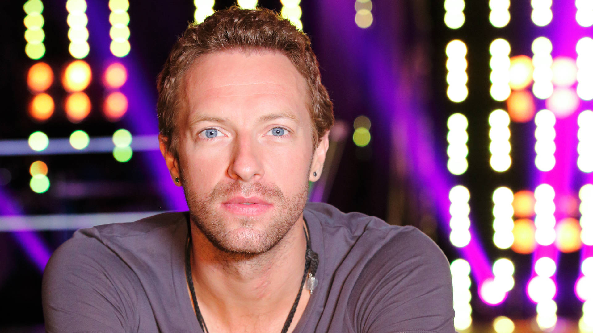 Chris Martin 舞台熱情演出 壁紙 — 一個藍眼睛、短棕髮的男人直視鏡頭，背景是色彩繽紛舞台燈光形成的充滿活力的散景。