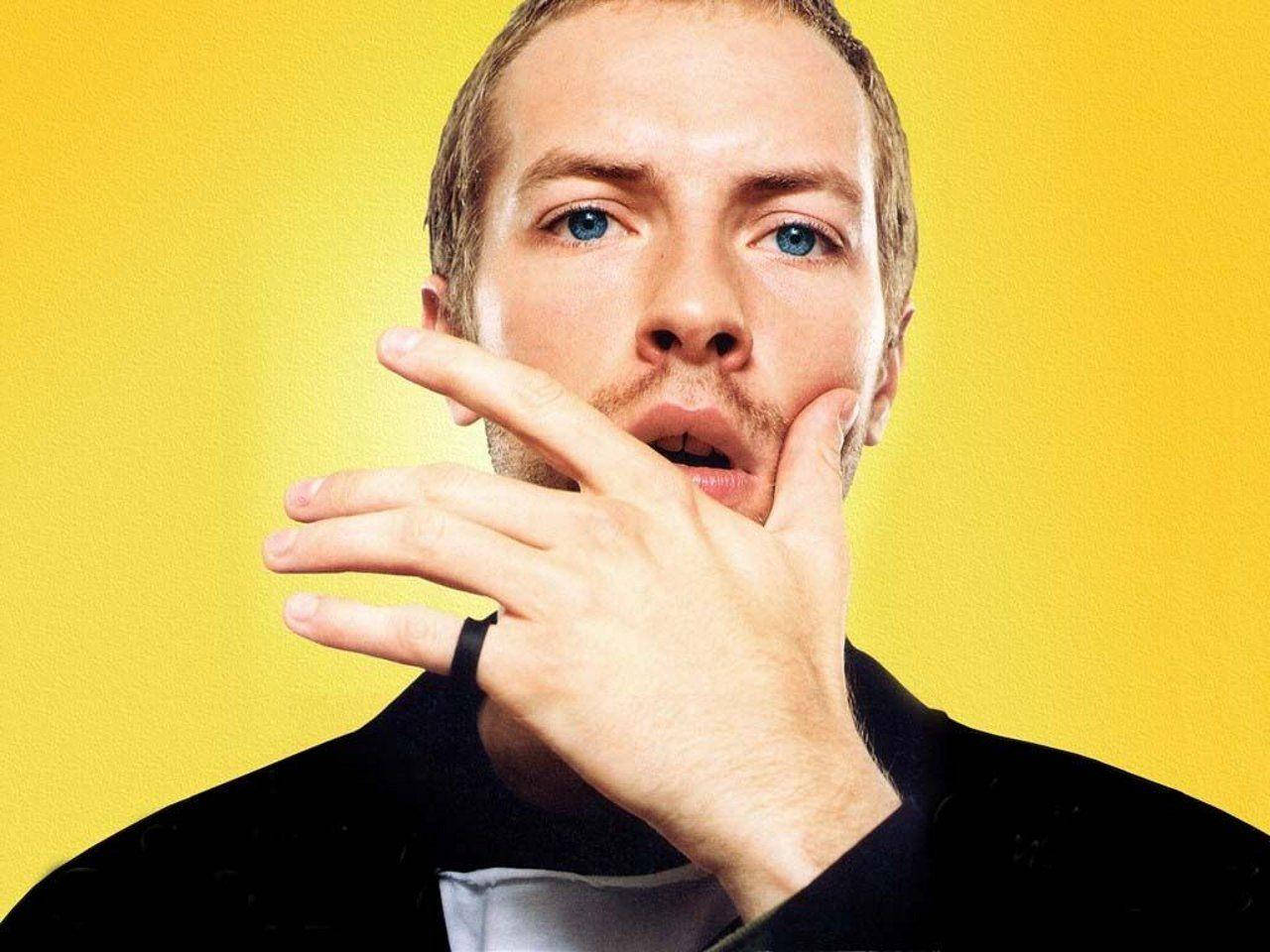 Chris Martin 黃色側影 壁紙 — 一個藍眼睛、留鬍鬚的男人，在明亮的黃色背景前將手靠近臉部。