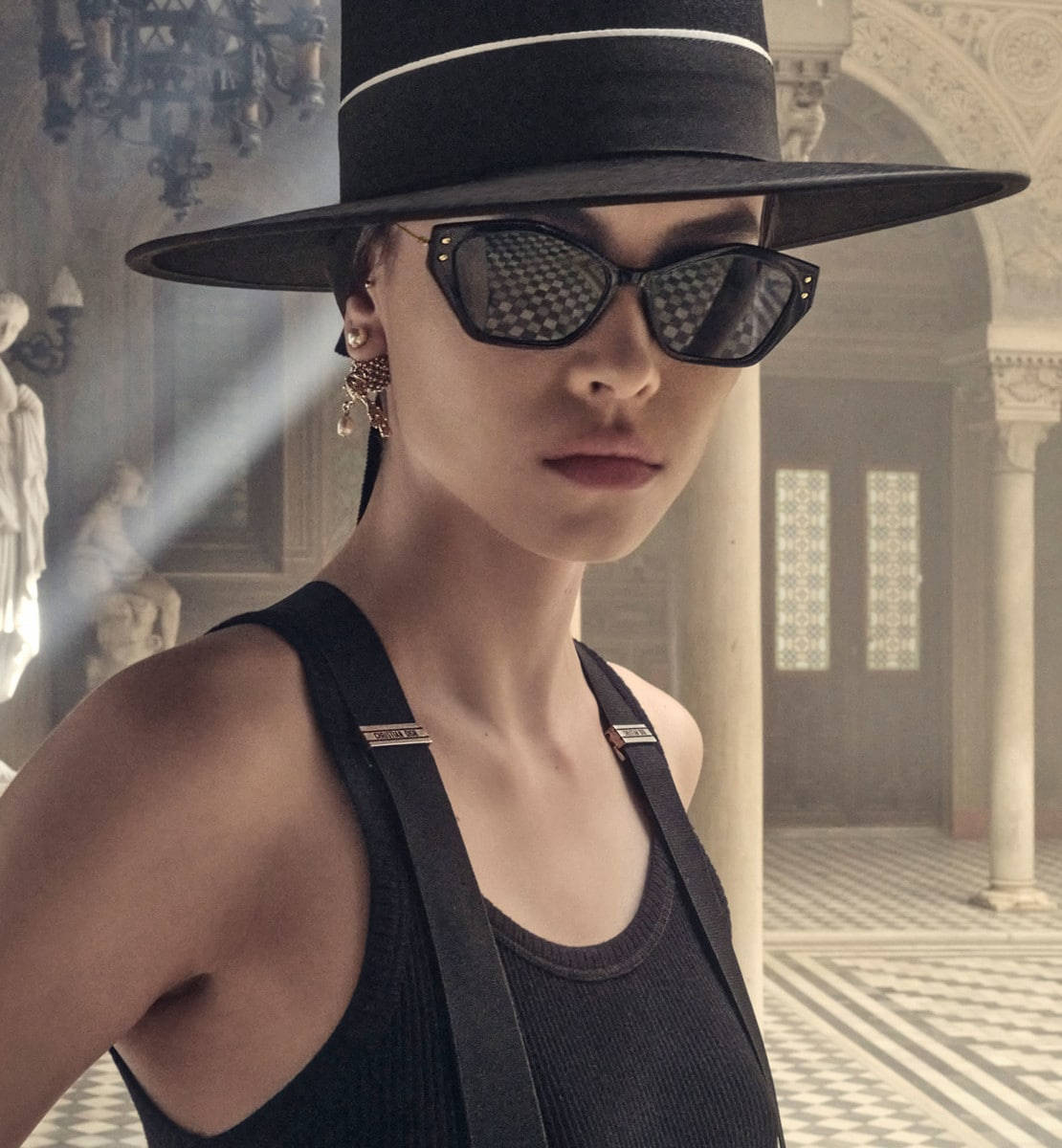 Christian Dior Miss Dior S1U太陽眼鏡 壁紙 — 一位戴著黑色帽子和太陽眼鏡的女性，站在一個陽光普照、裝飾華麗的大廳中，大廳有大理石柱和雕像。