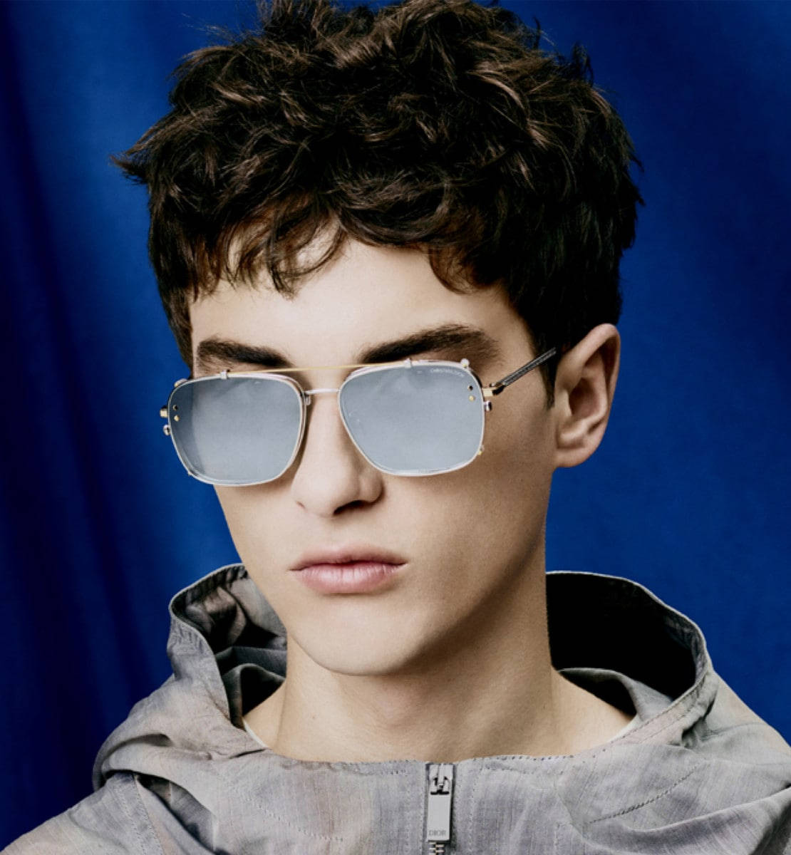Christian Dior 精緻銀色太陽眼鏡 壁紙 — 一位頭髮凌亂的深色頭髮年輕男子，在深藍色攝影棚背景下戴著反光飛行員太陽眼鏡。