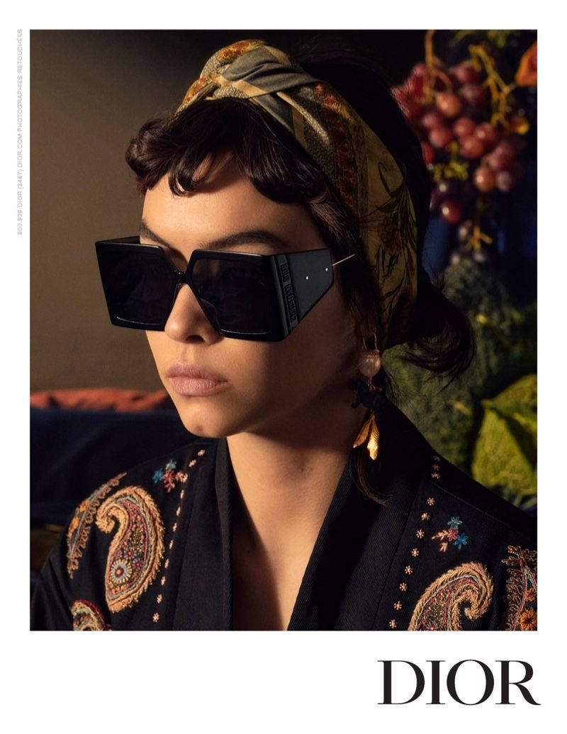 Christian Dior 高質感太陽眼鏡 壁紙 — 一位深色頭髮、戴著大黑色太陽眼鏡的女性，在葡萄和樹葉的背景前，戴著圖案頭巾和華麗的刺繡外套。