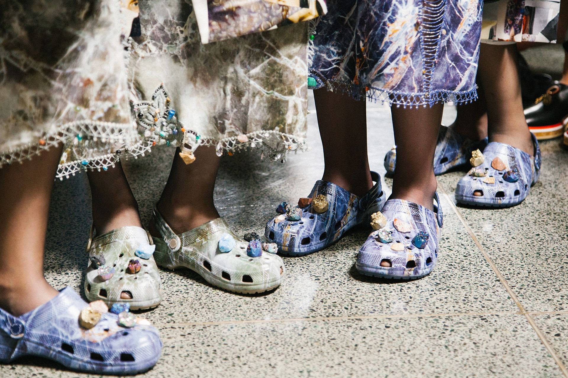 Christopher Kane X Crocs 聯名款 壁紙 — 兒童腳部特寫，穿著色彩繽紛、帶有珠飾的 Crocs 鞋，站在斑點地板瓷磚上。