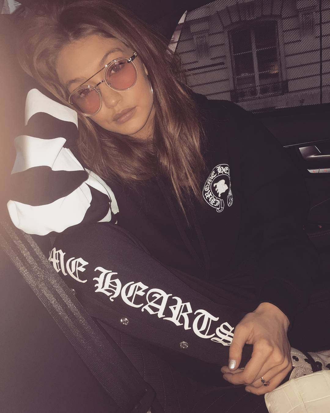 身穿 Chrome Hearts 套裝的模特 壁紙 — 一位長髮女子戴著粉紅色太陽眼鏡，身穿帶有白色字樣和骷髏圖案的黑色外套，在車內休息。