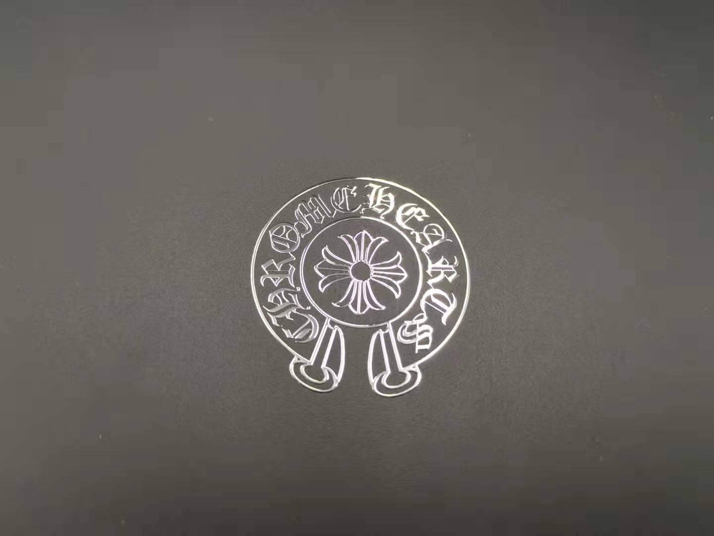 Chrome Hearts 貼紙貼紙 壁紙 — 一個銀色圓形徽章，帶有華麗的哥德式字體和中央花卉圖案，放置在深灰色紋理表面上。