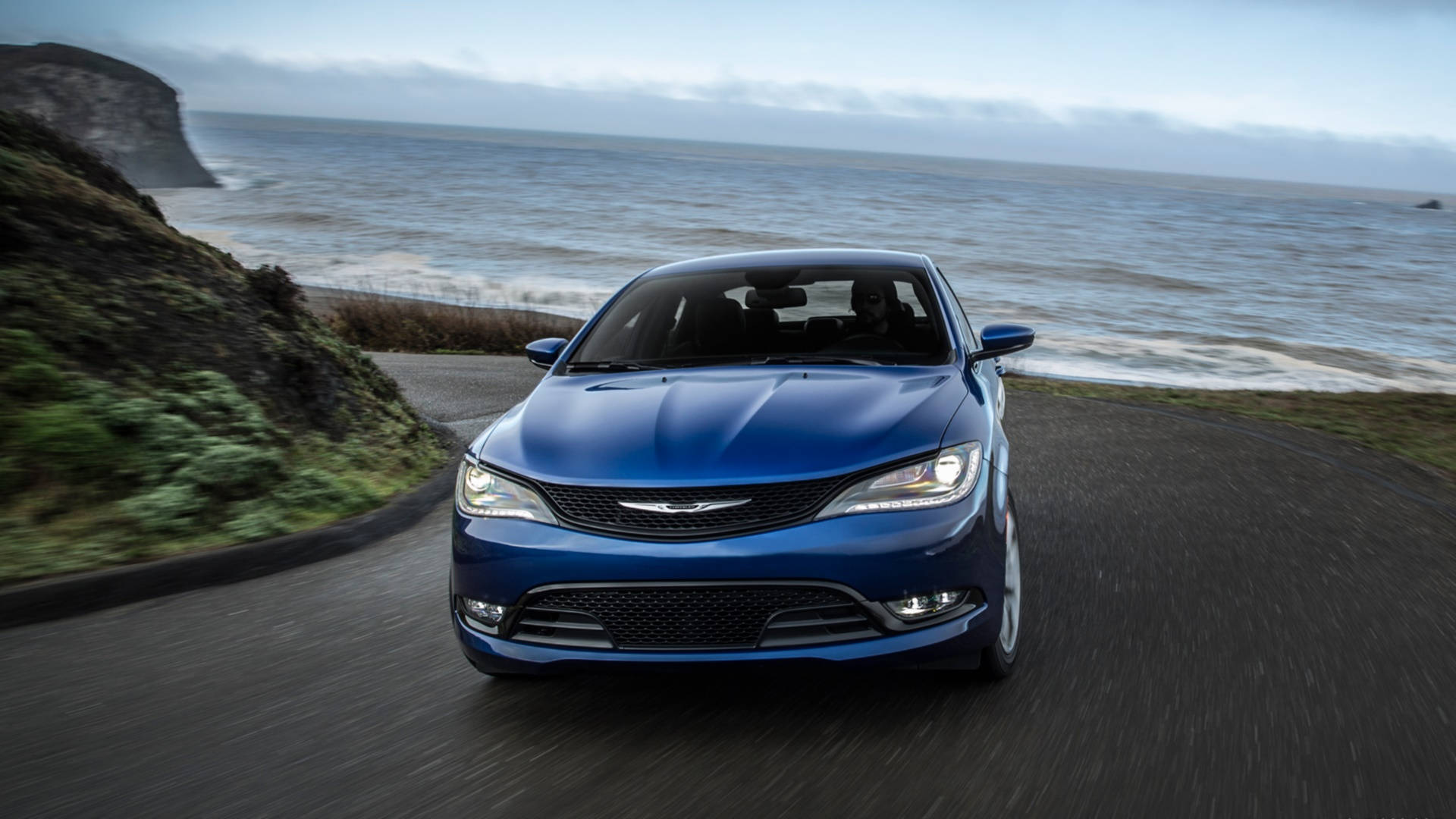 藍色 Chrysler 200 S 壁紙 — 一架時尚的藍色轎車沿著海岸公路飛馳，海浪拍打懸崖，天空陰沉。