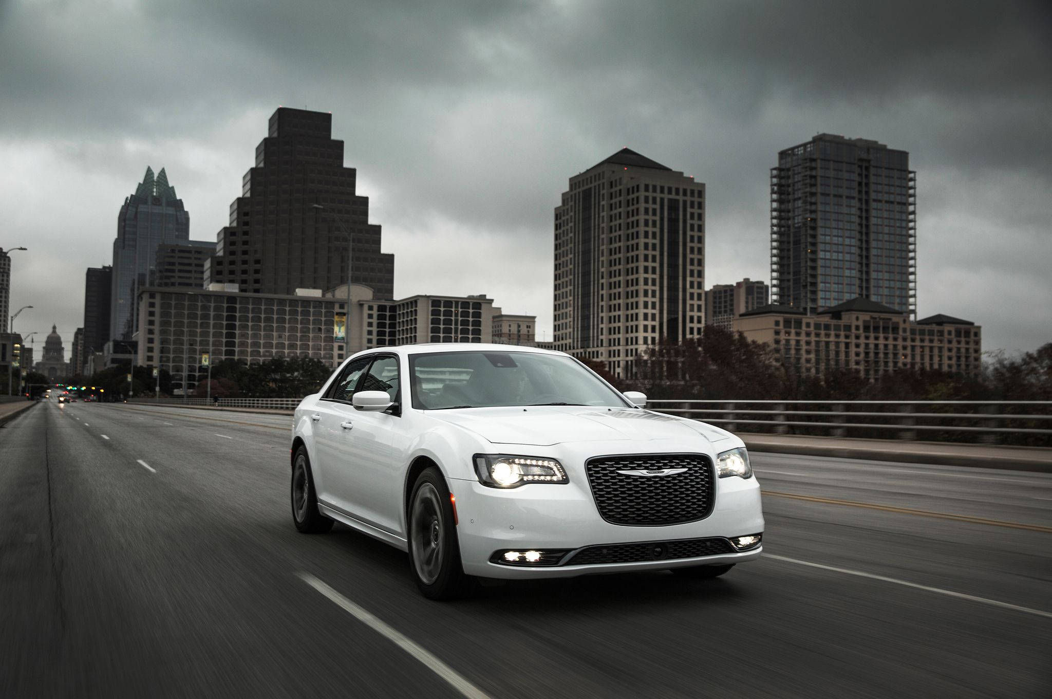 白色 Chrysler 300 壁紙 — 一輛白色豪華轎車在陰天之下疾馳過橋，背景是現代城市天際線。