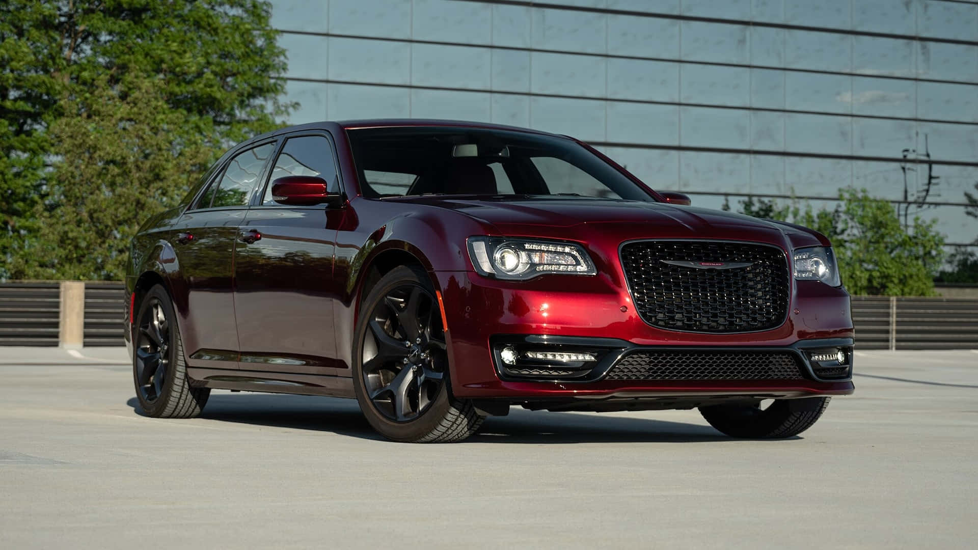 Chrysler 火力全開，釋放性能本質 壁紙 — 一輛光澤的栗色Chrysler 300轎車，配黑色輪圈，停在混凝土停車場上，背景是玻璃建築和綠樹。