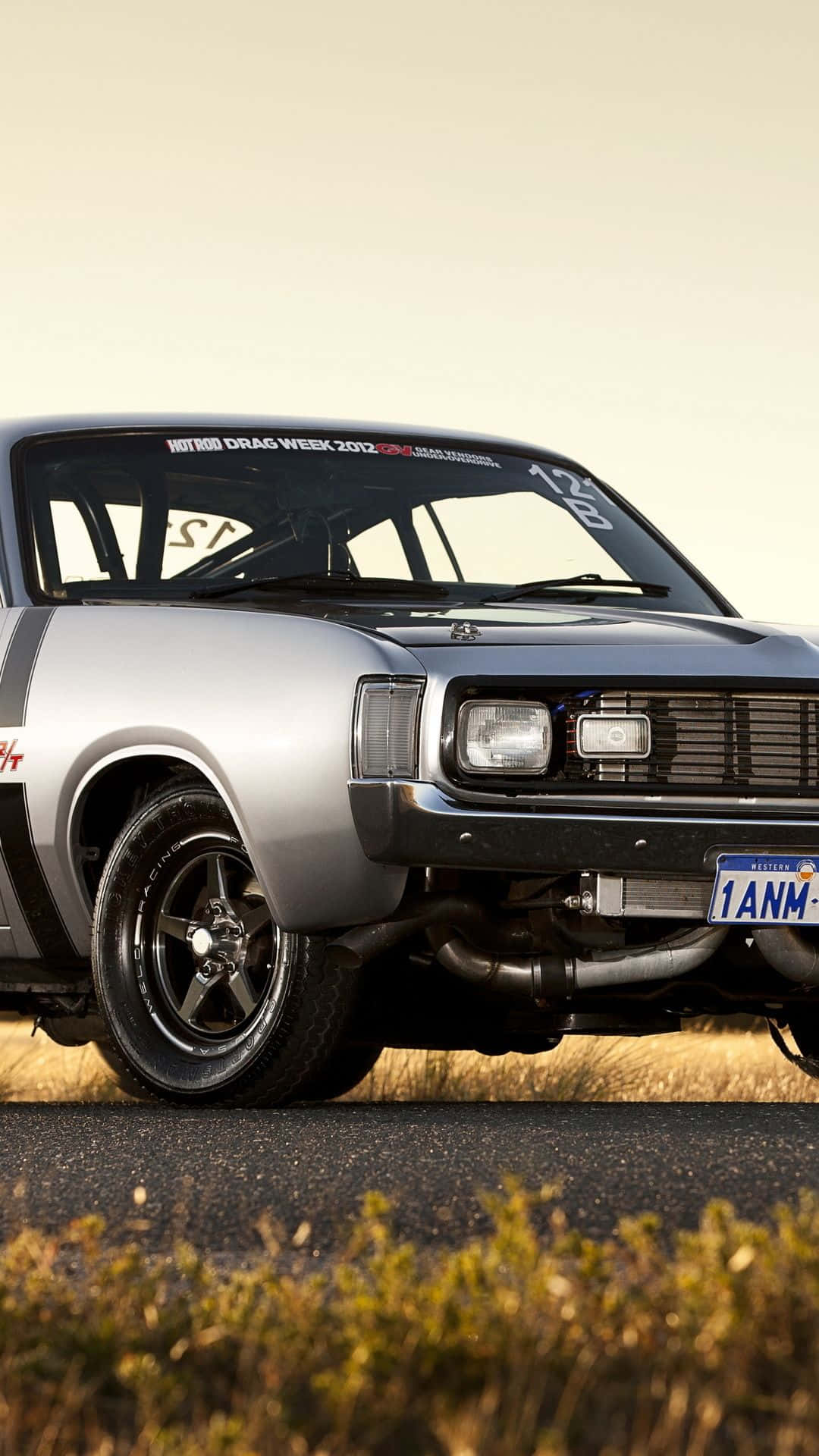 經典銀色 Chrysler Valiant Charger 跑車 iPhone 壁紙 — 一架銀色經典賽車，黑色車輪和暴露的排氣管，停在瀝青路上，旁邊是乾草，天空朦朧。