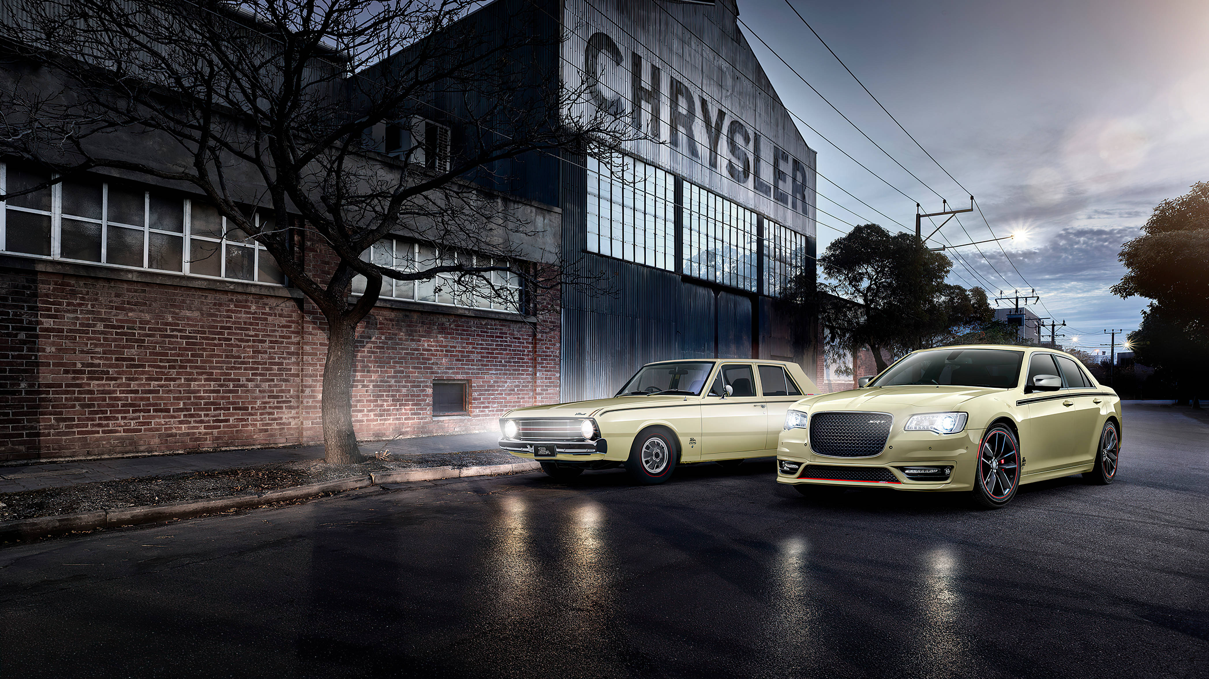 Chrysler 車輛藝術圖 壁紙 — 兩輛淡黃色克萊斯勒轎車在暮色燈光下閃閃發光，旁邊是一座歷史悠久的工廠，側面刻有「CHRYSLER」字樣。