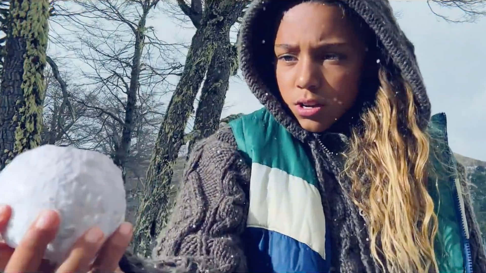  壁紙 — 一個穿著連帽冬季外套的女孩在戶外雪地森林中手持雪球，周圍是光禿禿的樹木。