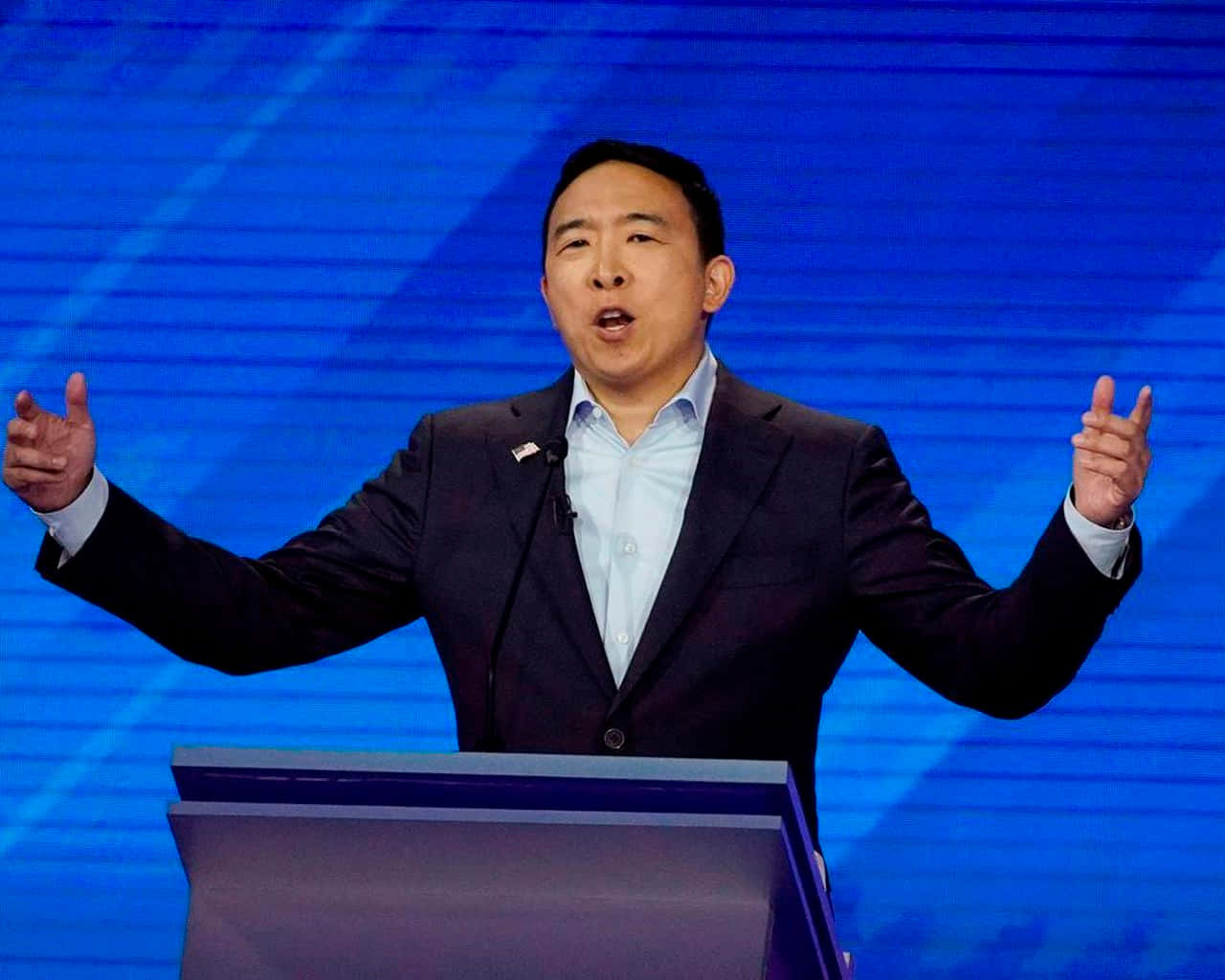 USA 桌布 壁紙 — 一名身穿深色西裝的男子站在講台上，對著充滿活力的藍色條紋背景熱情地做手勢。