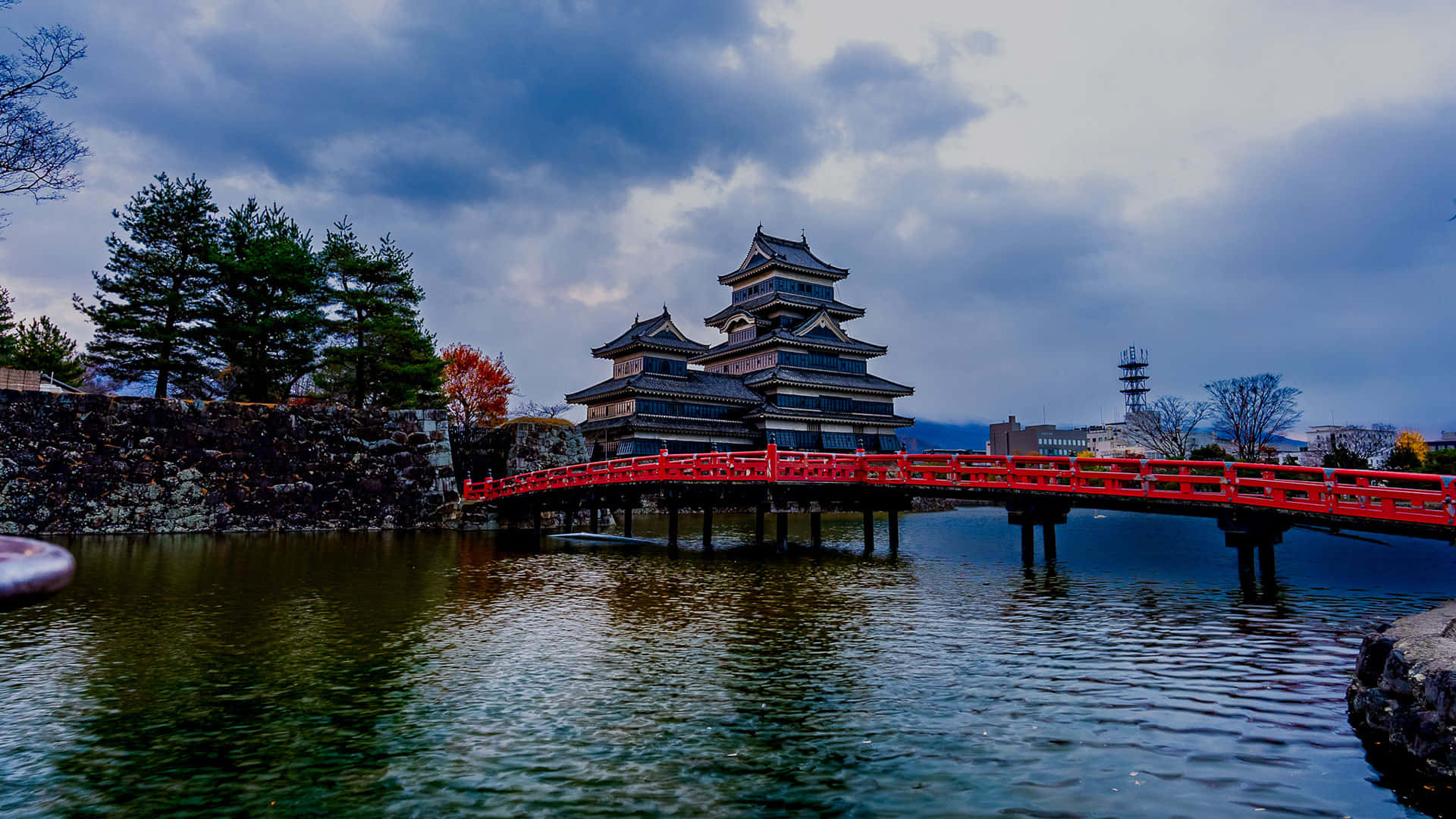  壁紙 — 一座傳統日本城堡，擁有層疊屋頂，矗立在一座鮮紅色橋樑後方，橋樑橫跨平靜水面，上方是陰沉多雲的天空。