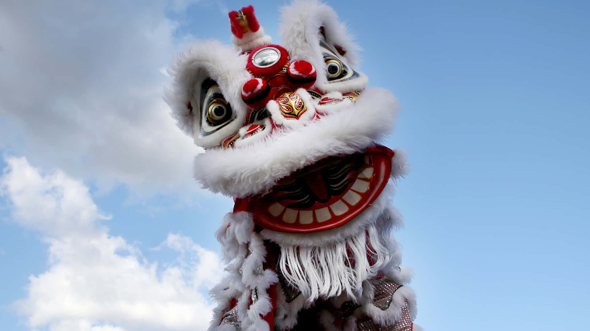  壁紙 — 一件鮮豔的紅白傳統中國舞獅服裝，帶有蓬鬆的白色毛皮和精緻的刺繡，矗立在晴朗的藍天白雲下。