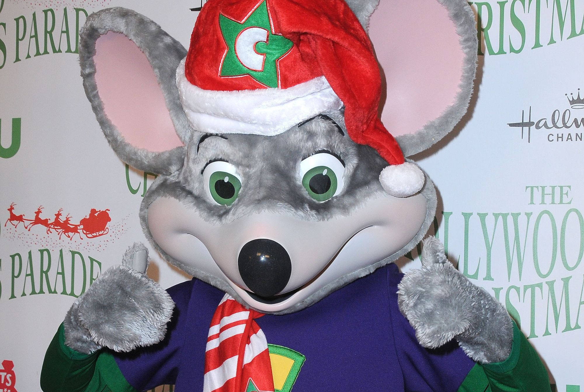 Chuck E Cheese聖誕遊行 壁紙 — 一隻快樂的灰色老鼠吉祥物，戴著紅色聖誕帽和條紋圍巾，為節日活動擺姿勢。
