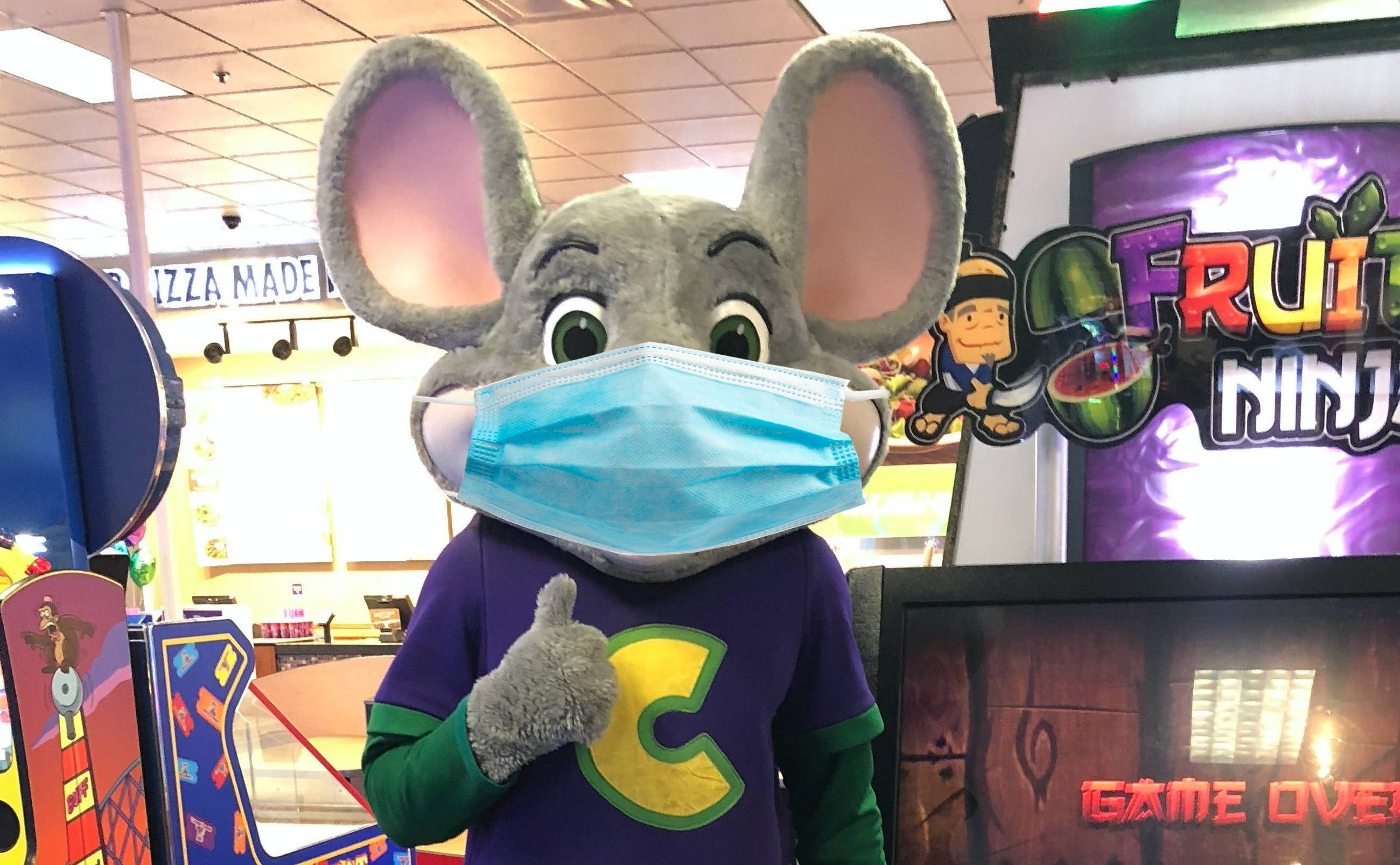 戴著口罩的 Chuck E Cheese 機械人 壁紙 — 一隻灰色老鼠吉祥物戴著藍色外科口罩，在光線明亮的遊戲機中心豎起大拇指，身後有遊戲機。