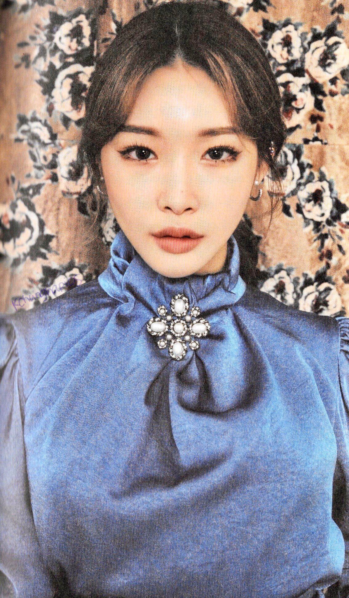 Chungha 藍色禮服 壁紙 — 一位身穿藍色高領襯衫、佩戴鑲寶石胸針的女性，在花卉壁紙背景下向前凝視。
