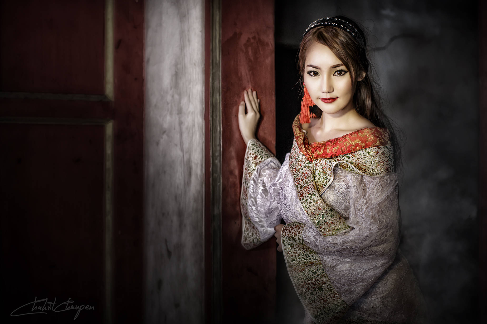 穿傳統泰國「Chut Thai」服飾的美女 壁紙 — 一位身穿華麗傳統服飾的女性，在昏暗且充滿氛圍的環境中，從一扇風化的木門後探出頭來。