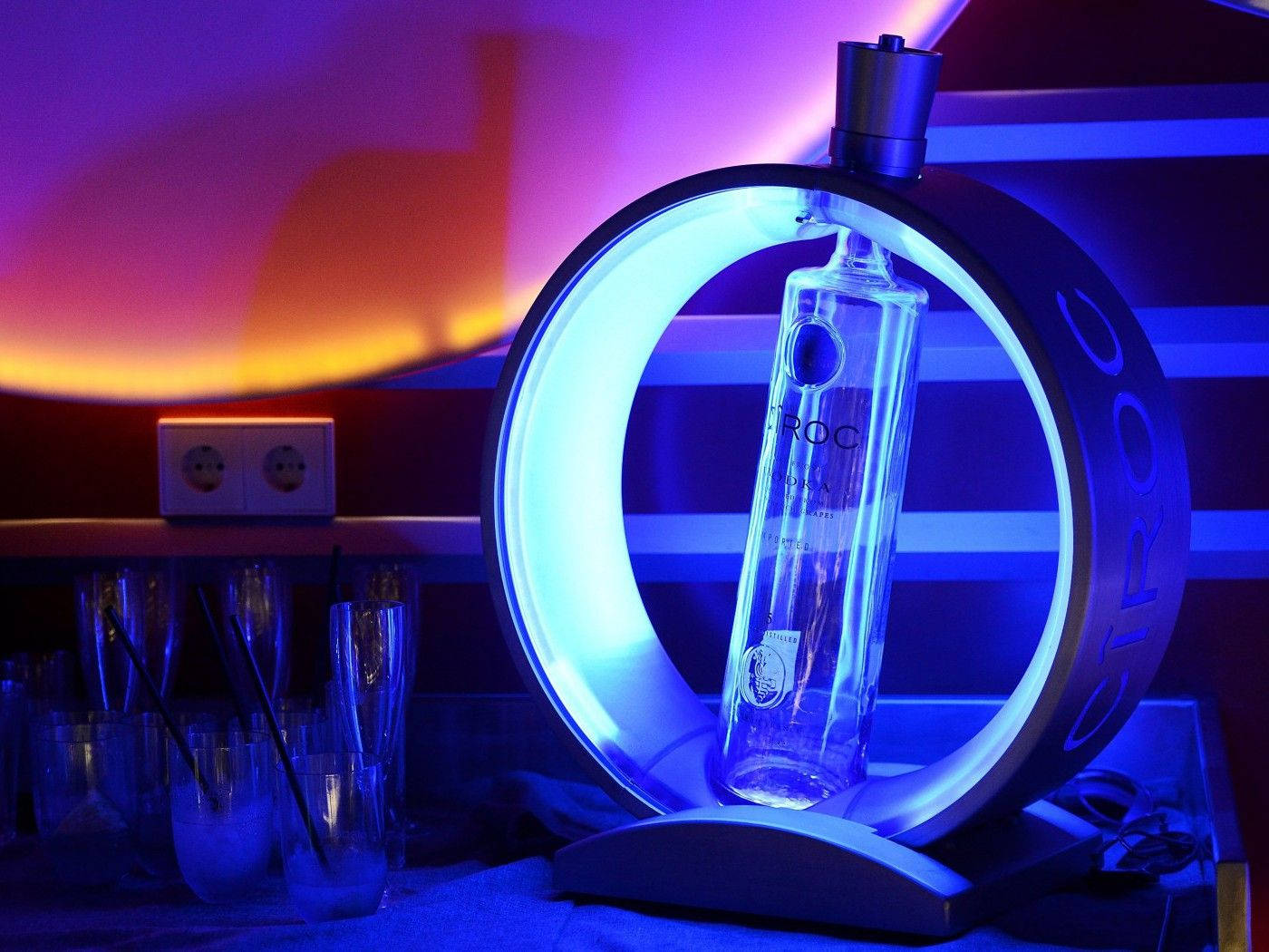 標語：優雅的 Ciroc 法國伏特加瓶身 壁紙 — 一個發光的藍色環形燈照亮了一個標有 'Ciroc' 品牌的透明伏特加酒瓶，放在一個時尚的支架上，背景是充滿活力的紫色和橙色漸變牆。