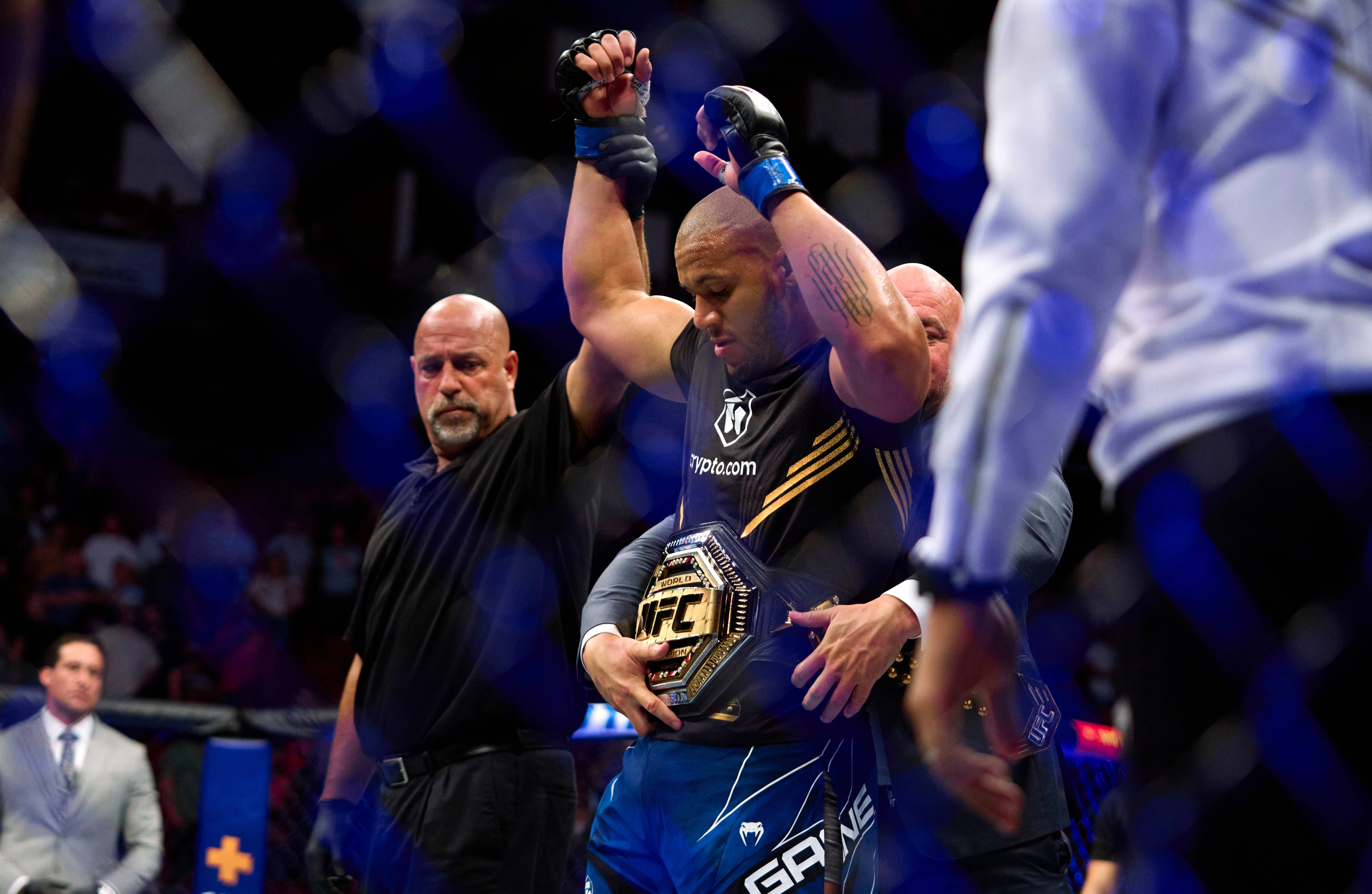 Ciryl Gane 被宣布為勝者 壁紙 — 一位勝利的UFC拳手在明亮的場館燈光下，於八角籠中高舉冠軍腰帶和手臂，裁判宣佈他為勝者。
