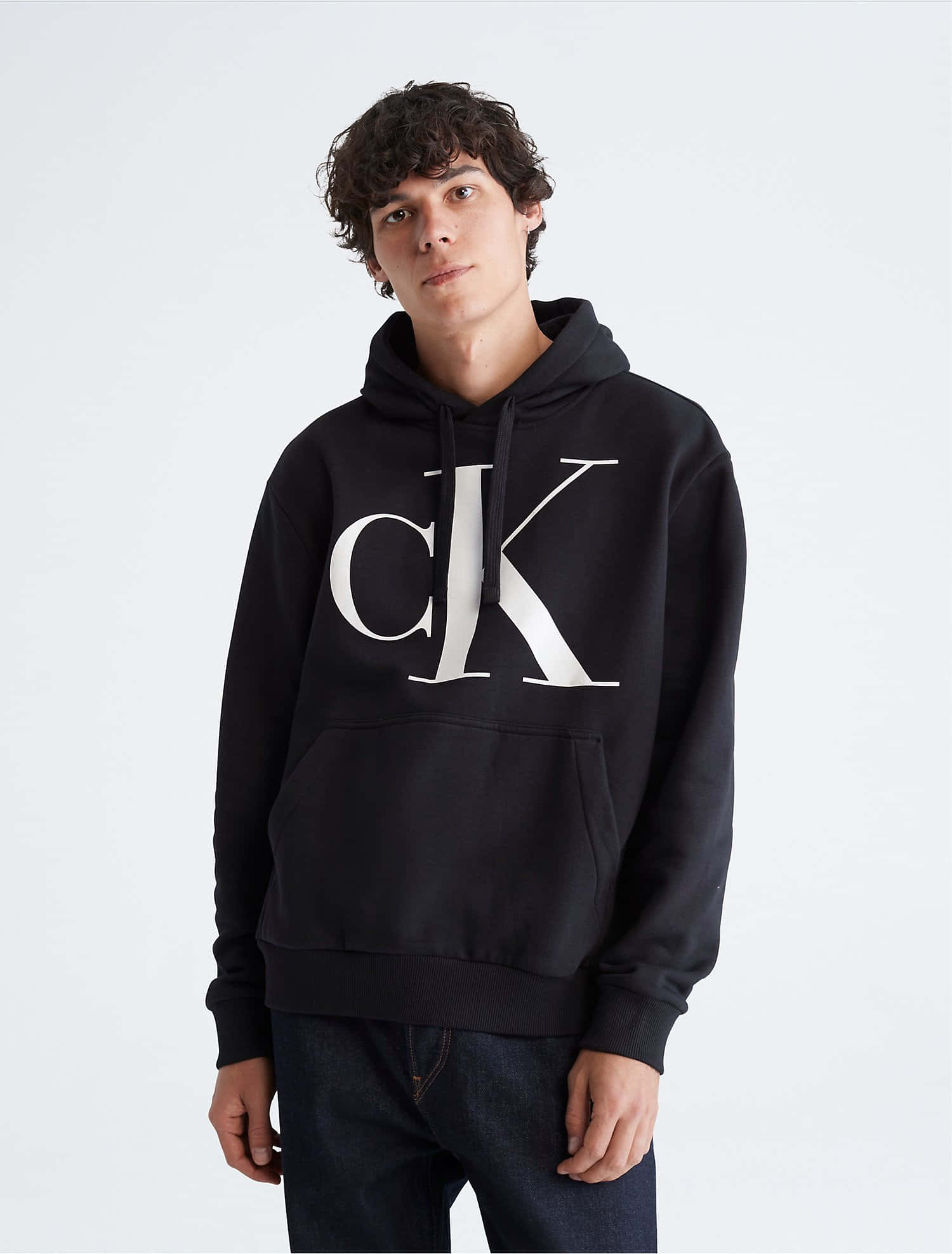 以CK展現你的自信 壁紙 — 一位年輕男子，留著黑色捲髮，身穿黑色Calvin Klein連帽衫，胸前有大型白色標誌，背景為純白色。