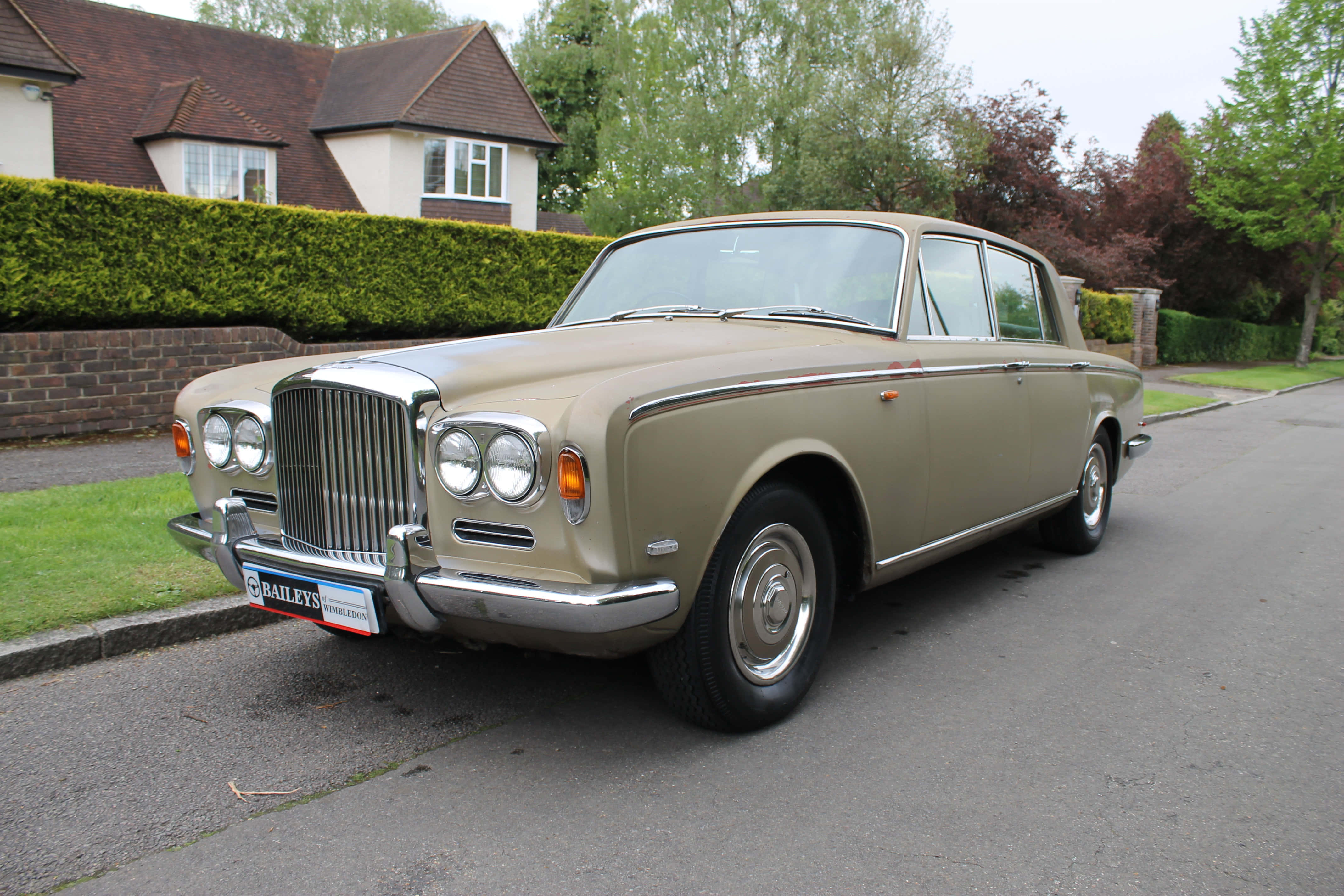 Bentley T1 桌布 壁紙 — 一輛經典米色賓利S1房車停泊在寧靜的住宅街道上，背景有綠色籬笆和磚屋。