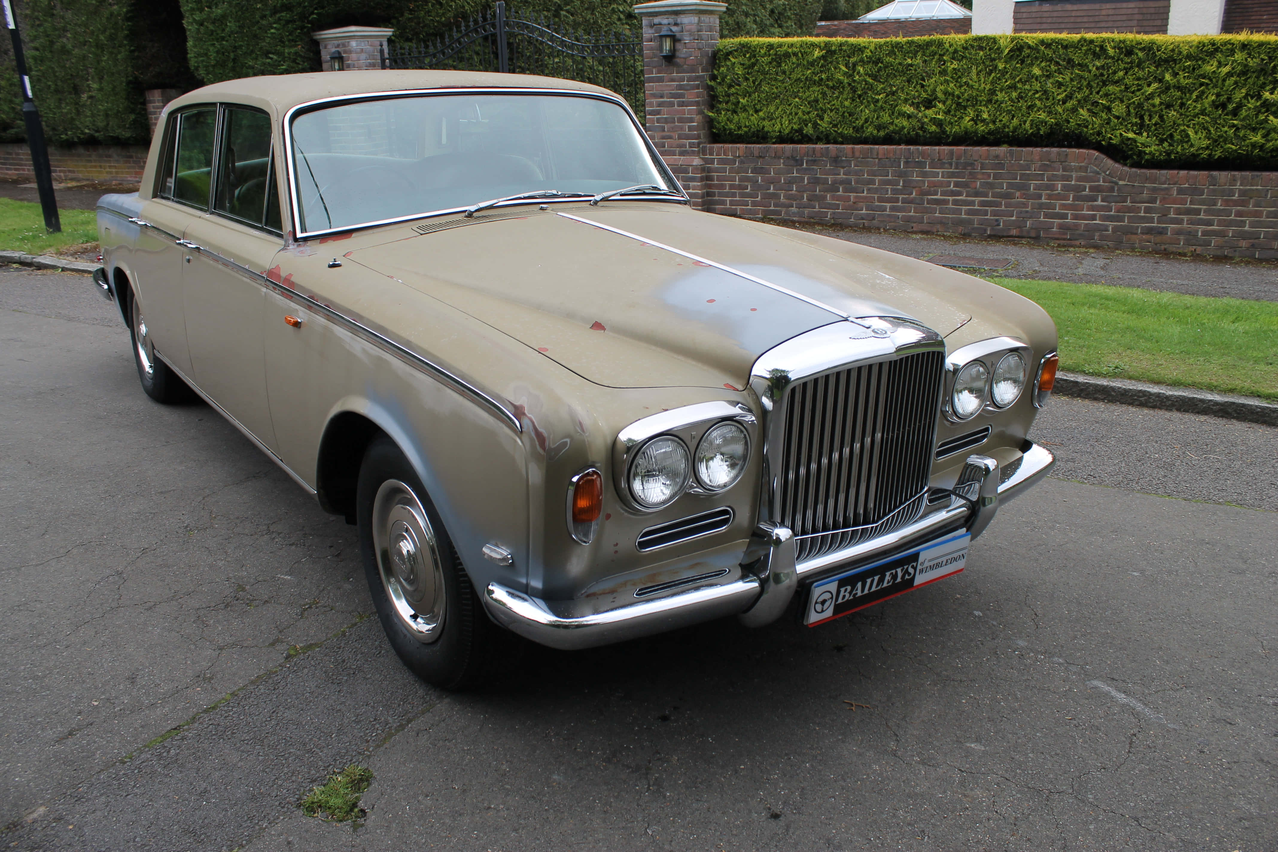 Bentley T1 桌布 壁紙 — 一輛經典米色賓利轎車，帶有鍍鉻裝飾，停在磚牆和綠色籬笆旁的鋪砌車道上。