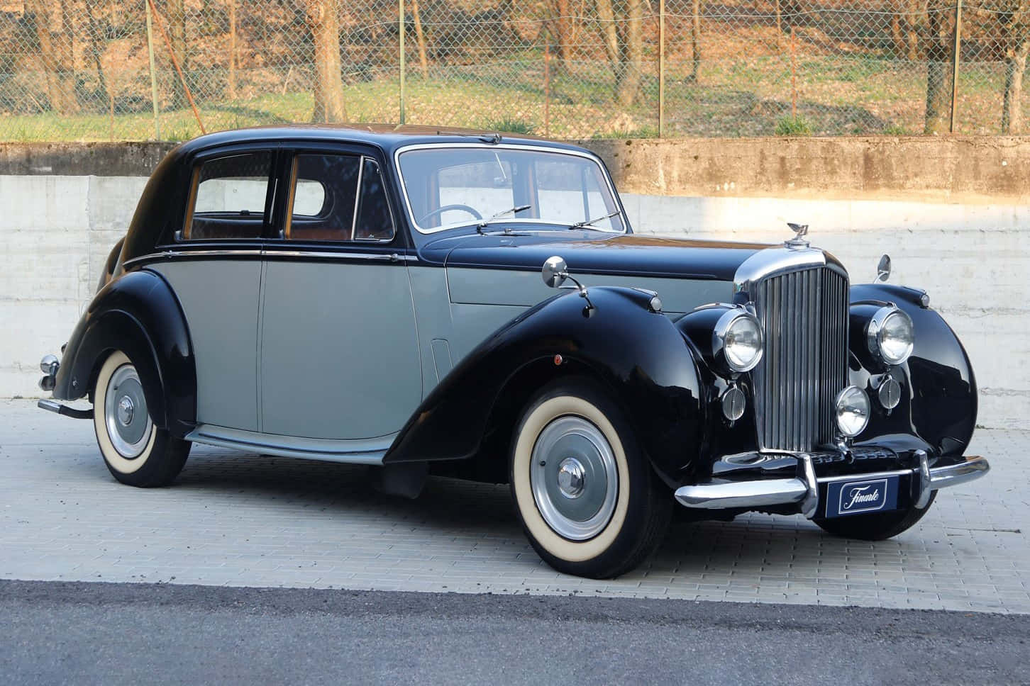 Bentley Mark Vi 桌布 壁紙 — 一輛經典黑色和銀色積架房車，配備奶油色壁輪胎，停泊在靠近圍欄和樹木的鋪砌地面上。