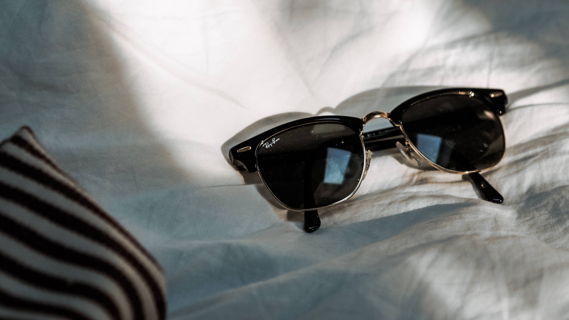 Ray-Ban Pinterest HD 壁紙 — 經典黑色太陽眼鏡配金色鏡框，放在柔和的自然光下皺巴巴的白色床單上。