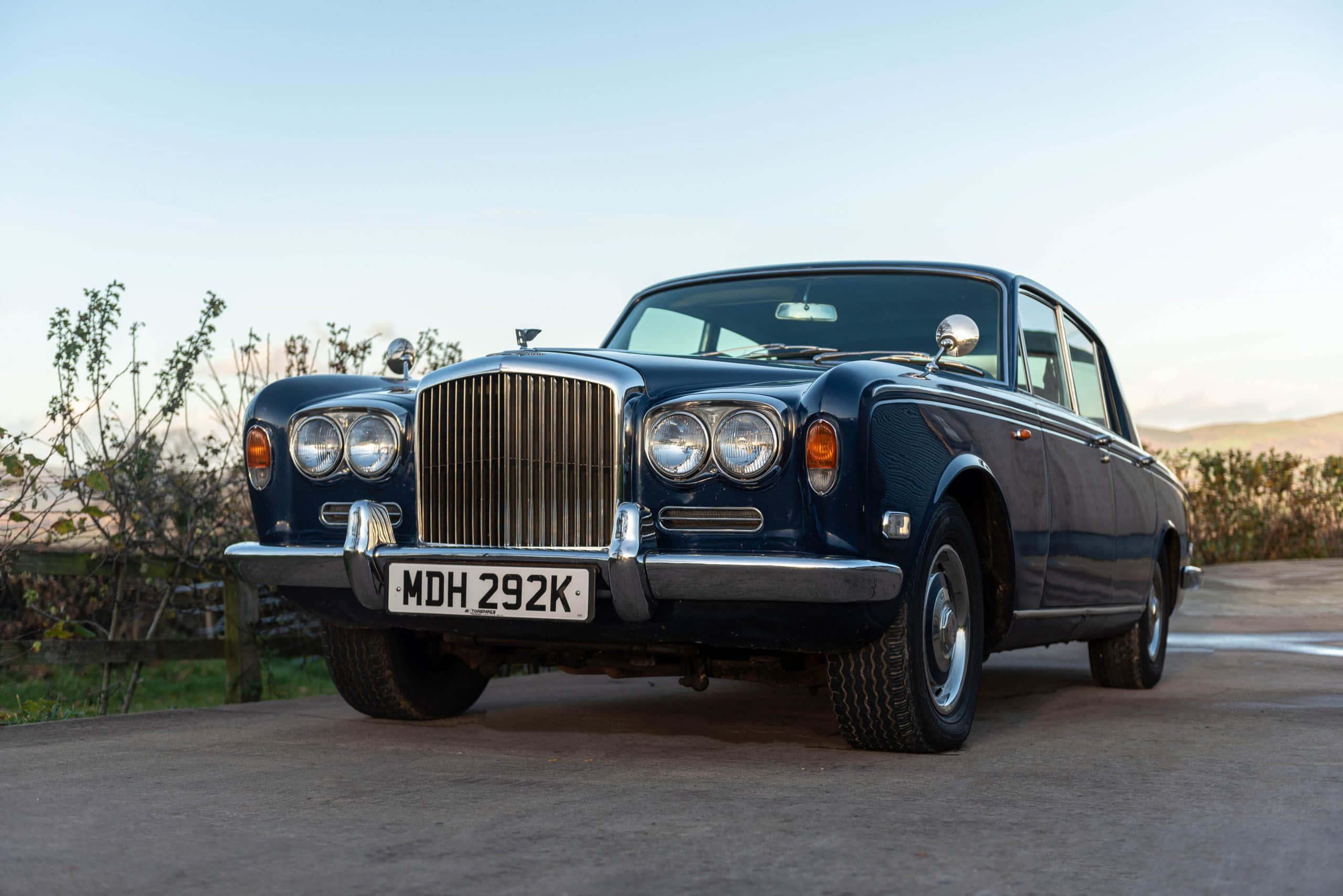 Bentley T1 桌布 壁紙 — 一輛經典藍色賓利S1轎車在寧靜的鄉村道路上，在柔和的金色時光下閃閃發光。