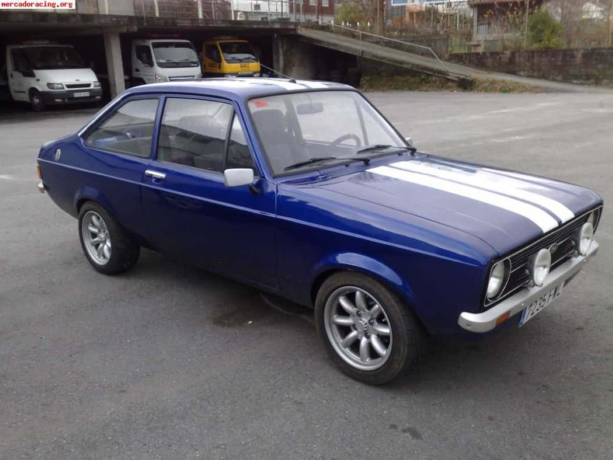 Ford Escort 桌布 壁紙 — 一輛經典藍色福特Escort，帶有白色賽車條紋，停放在陰天下的城市空地中。