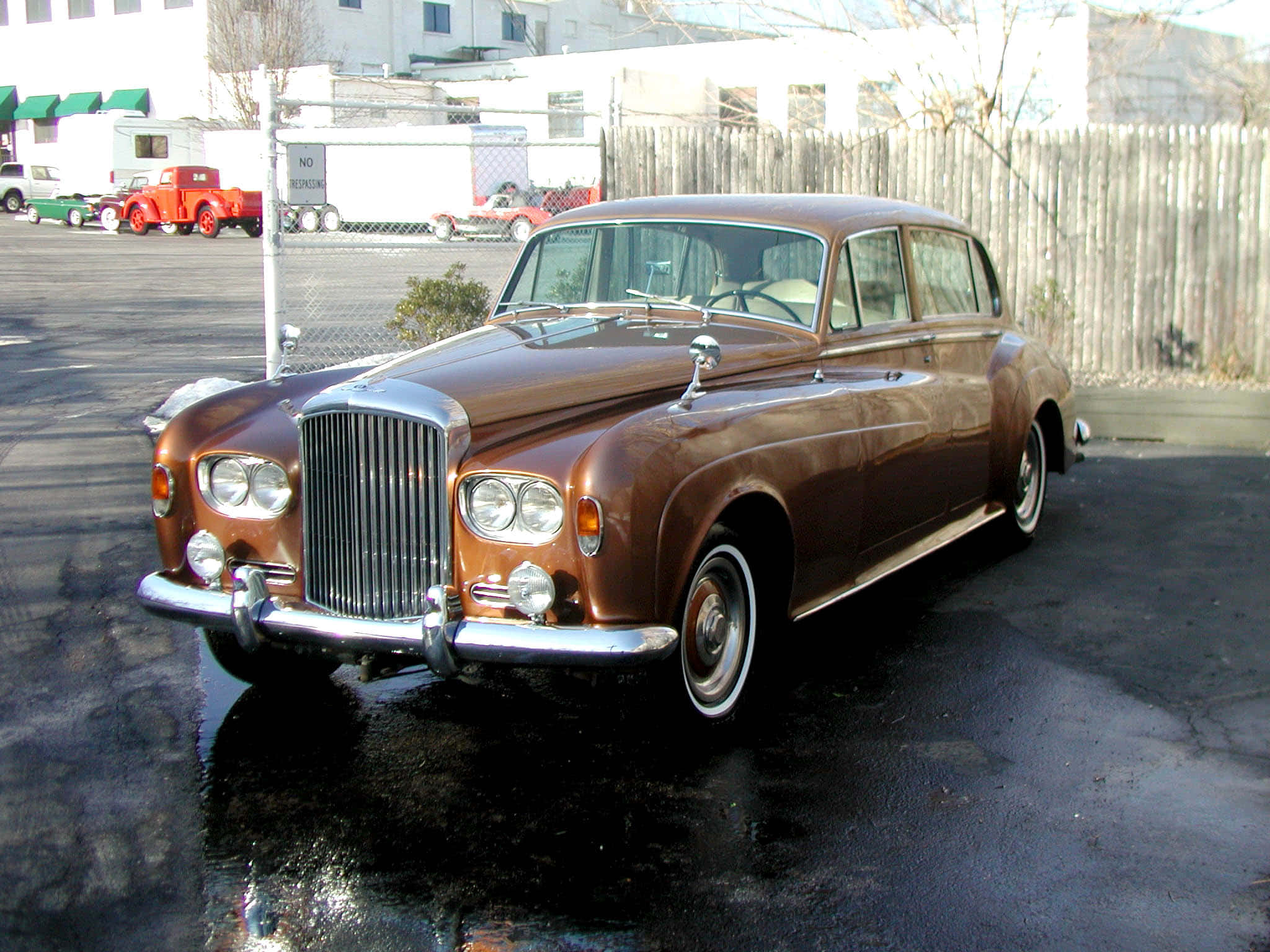Bentley S3 桌布 壁紙 — 一輛經典的棕色賓利轎車在明亮的日光下，在潮濕的路面上閃閃發光，背景有復古卡車。