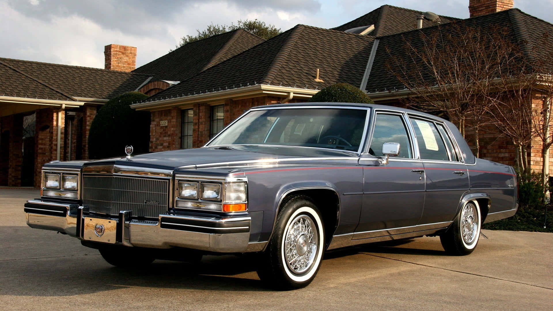 Cadillac Fleetwood 桌布 壁紙 — 一輛經典深灰色凱迪拉克轎車，帶有鍍鉻裝飾和白牆輪胎，停在磚房前的車道上。