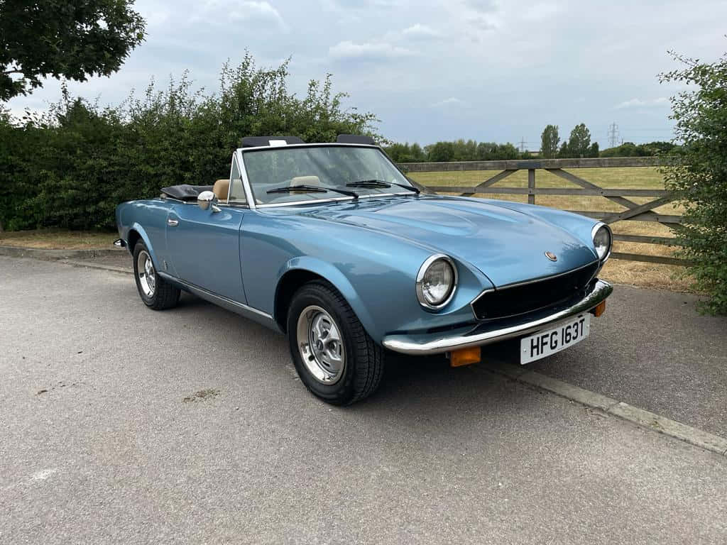 Fiat 124 Spider 桌布 壁紙 — 一輛經典淺藍色開篷跑車停泊在鋪砌道路上，旁邊有木柵欄和綠色灌木，天空多雲。