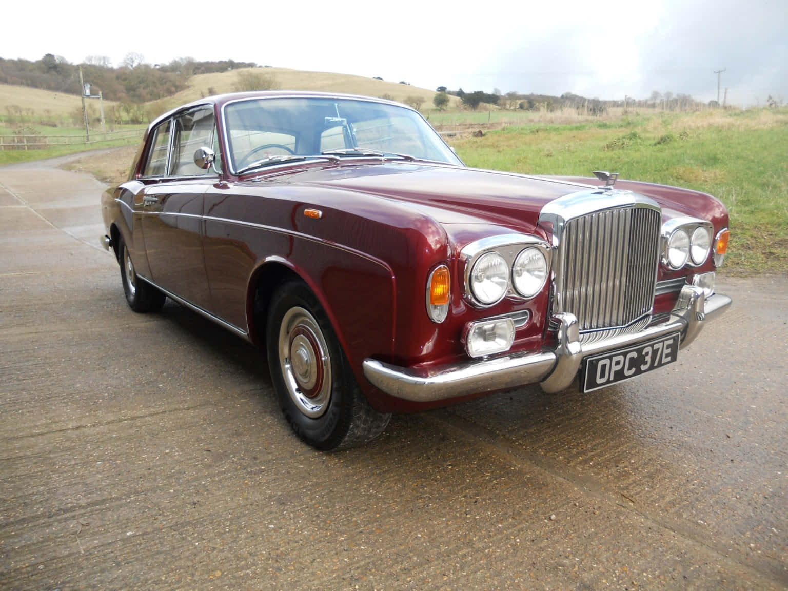 Bentley T1 桌布 壁紙 — 一輛經典的棗紅色賓利S2在陰天之下閃閃發光，停泊在濕潤的鄉村道路上，背景是連綿起伏的山丘。