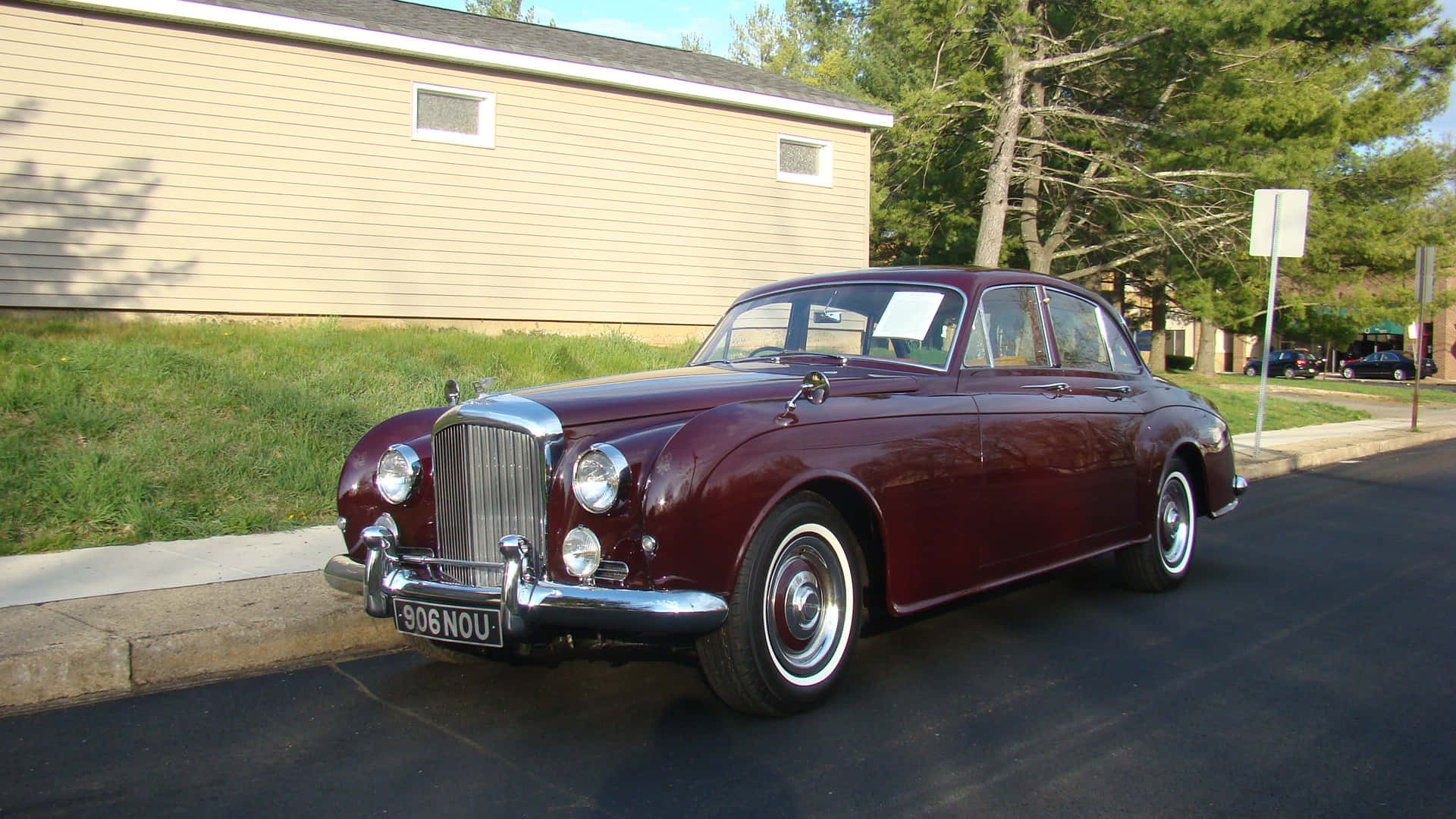 Bentley S2 桌布 壁紙 — 一輛經典的栗色賓利轎車在明亮的陽光下閃閃發光，停在郊區街道上，旁邊是米色房屋和綠色草坪。