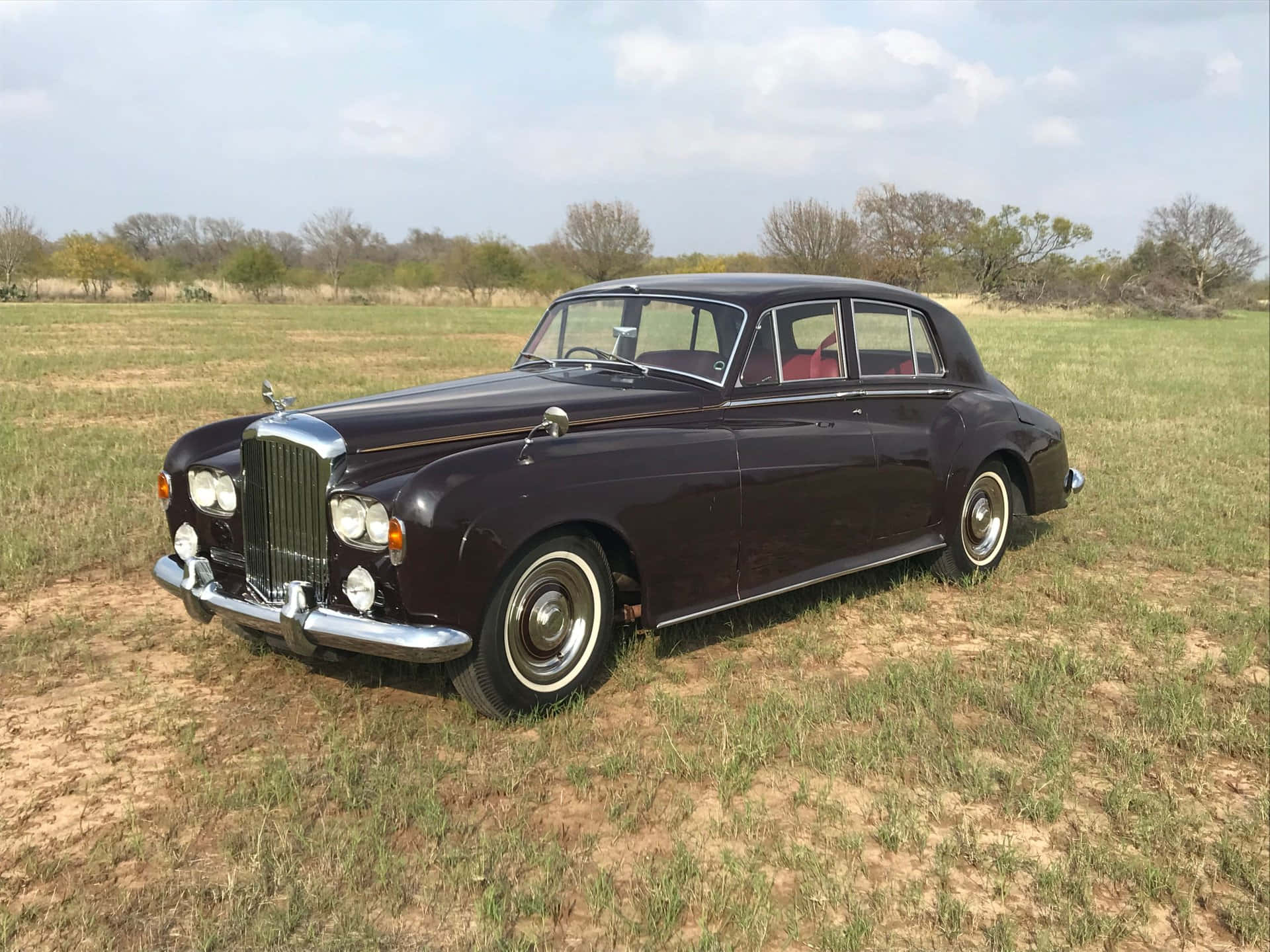 Bentley S3 桌布 壁紙 — 一輛經典的栗色賓利轎車，帶有鍍鉻裝飾，停放在陽光普照的草地上，天空部分多雲。