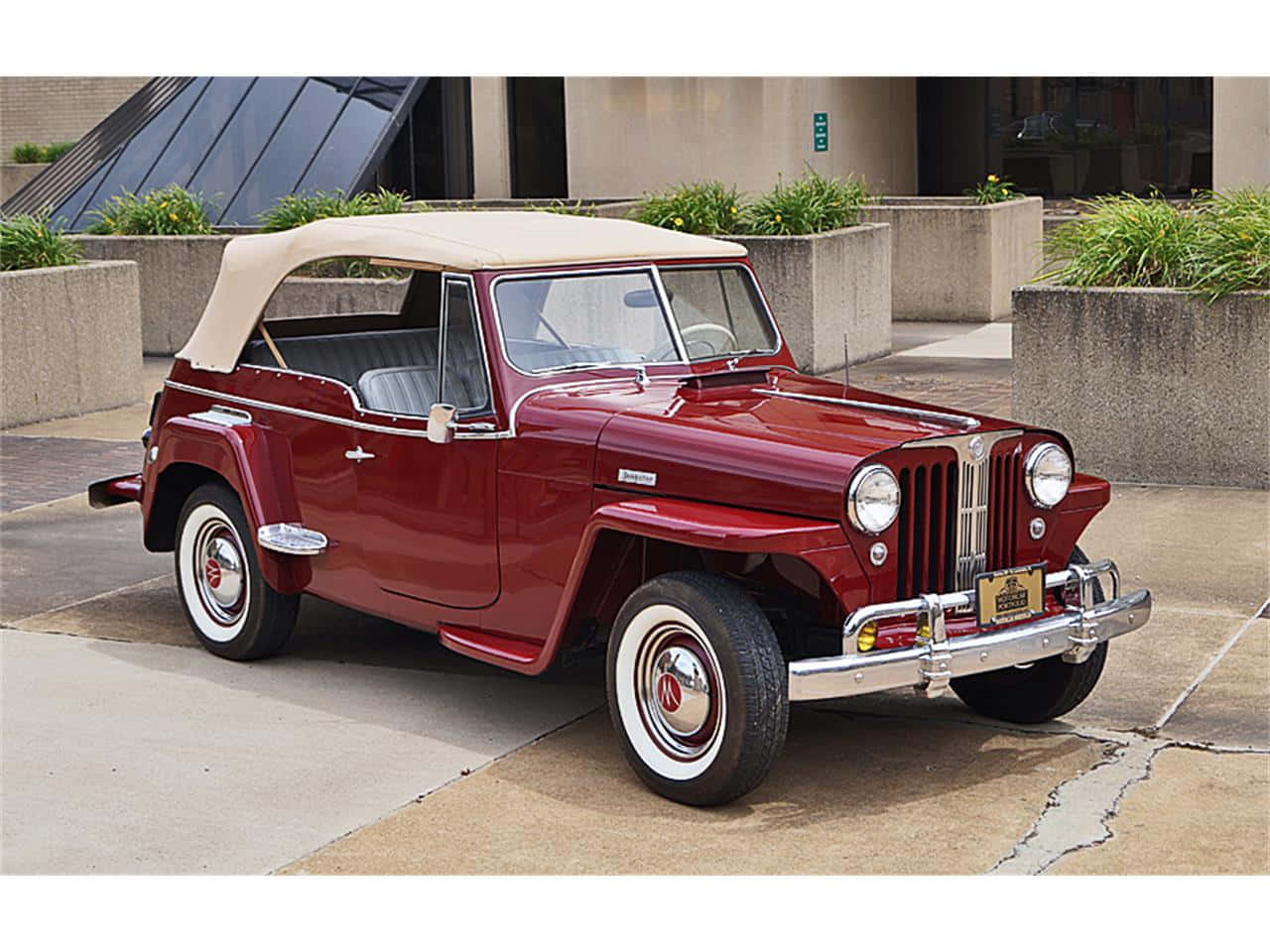 Jeepster 桌布 壁紙 — 經典栗色Jeepster開篷車配奶油色車頂，停泊在現代花盆環繞的混凝土廣場上。