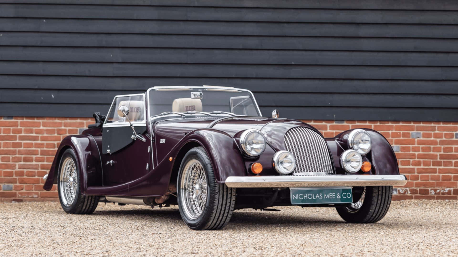 Morgan Roadster 桌布 壁紙 — 一輛經典栗色摩根跑車，帶有鍍鉻裝飾和鋼絲輪，停在磚牆和黑色木牆前的礫石上。